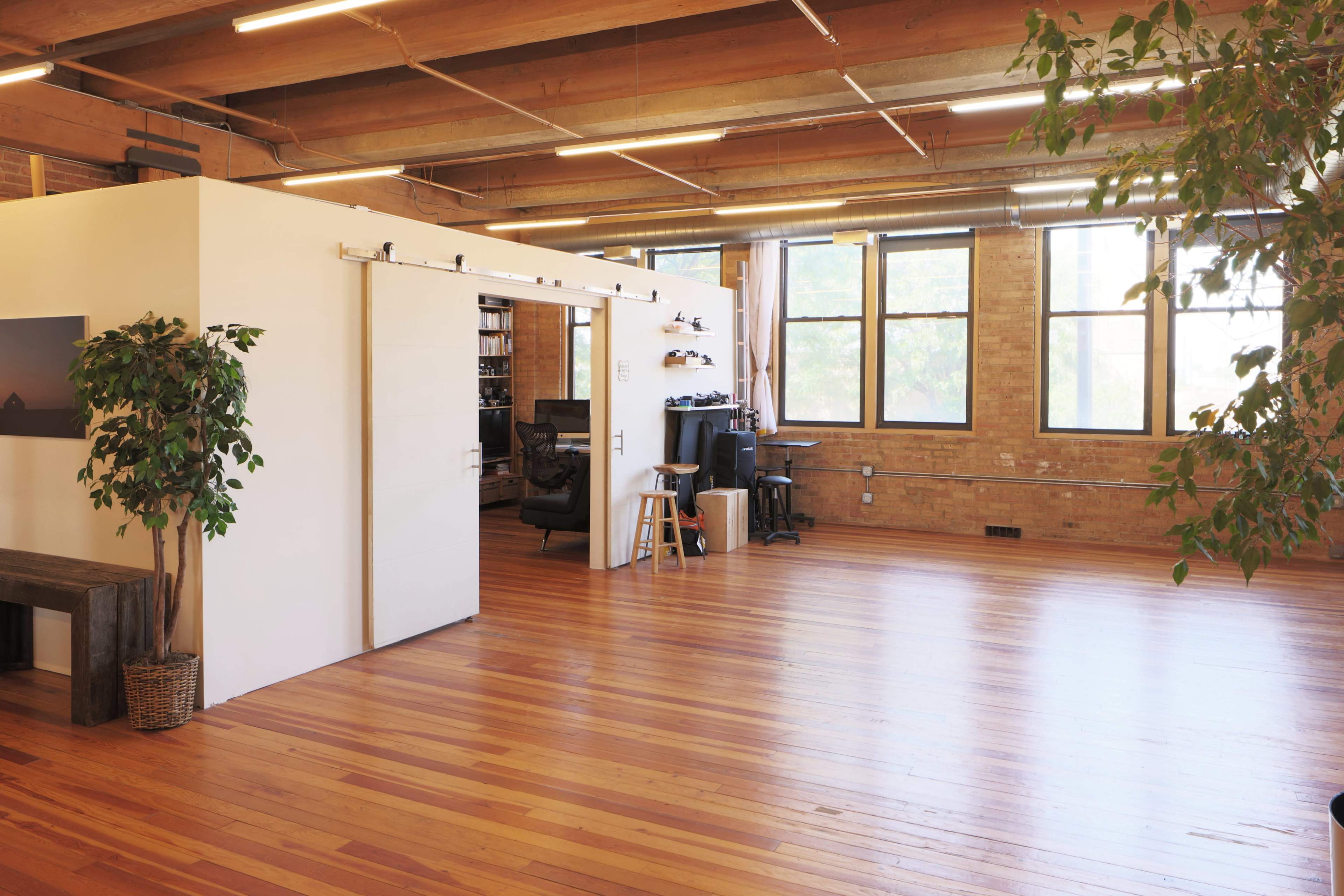The image shows a spacious, sunlit open office with wooden flooring, large windows, and a white partition separating two areas.