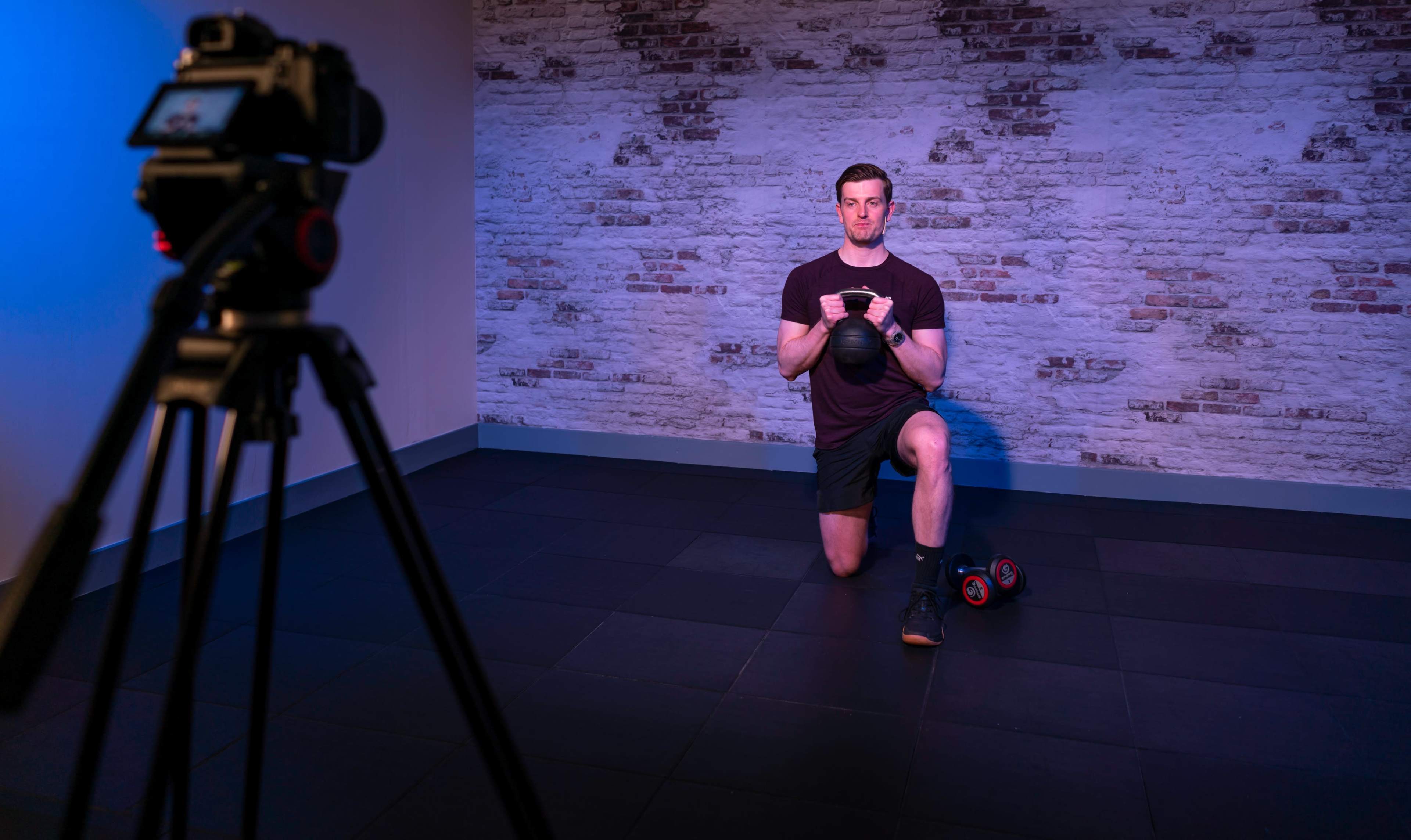 A man is kneeling on one leg, holding a kettlebell, while being filmed by a camera on a tripod in a studio with a brick wall.