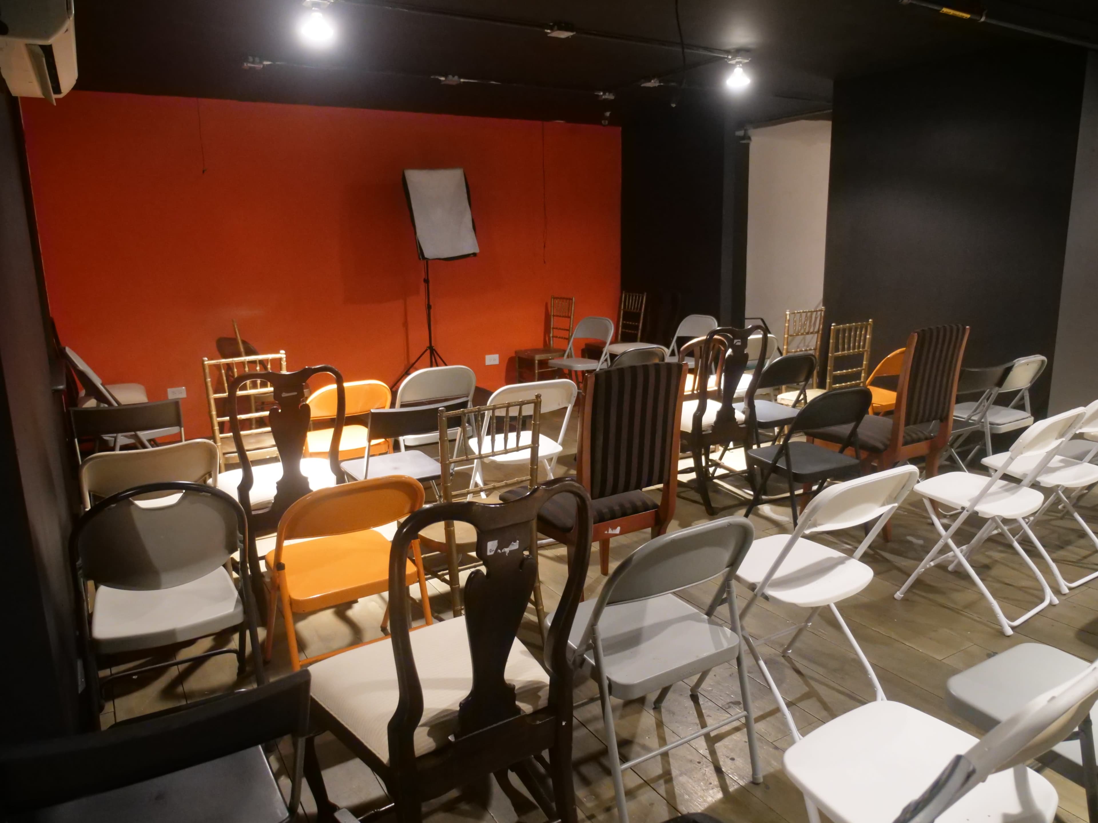 A room is set up with a variety of chairs arranged in rows on a wooden floor, with an orange wall and a blank stand in the background.