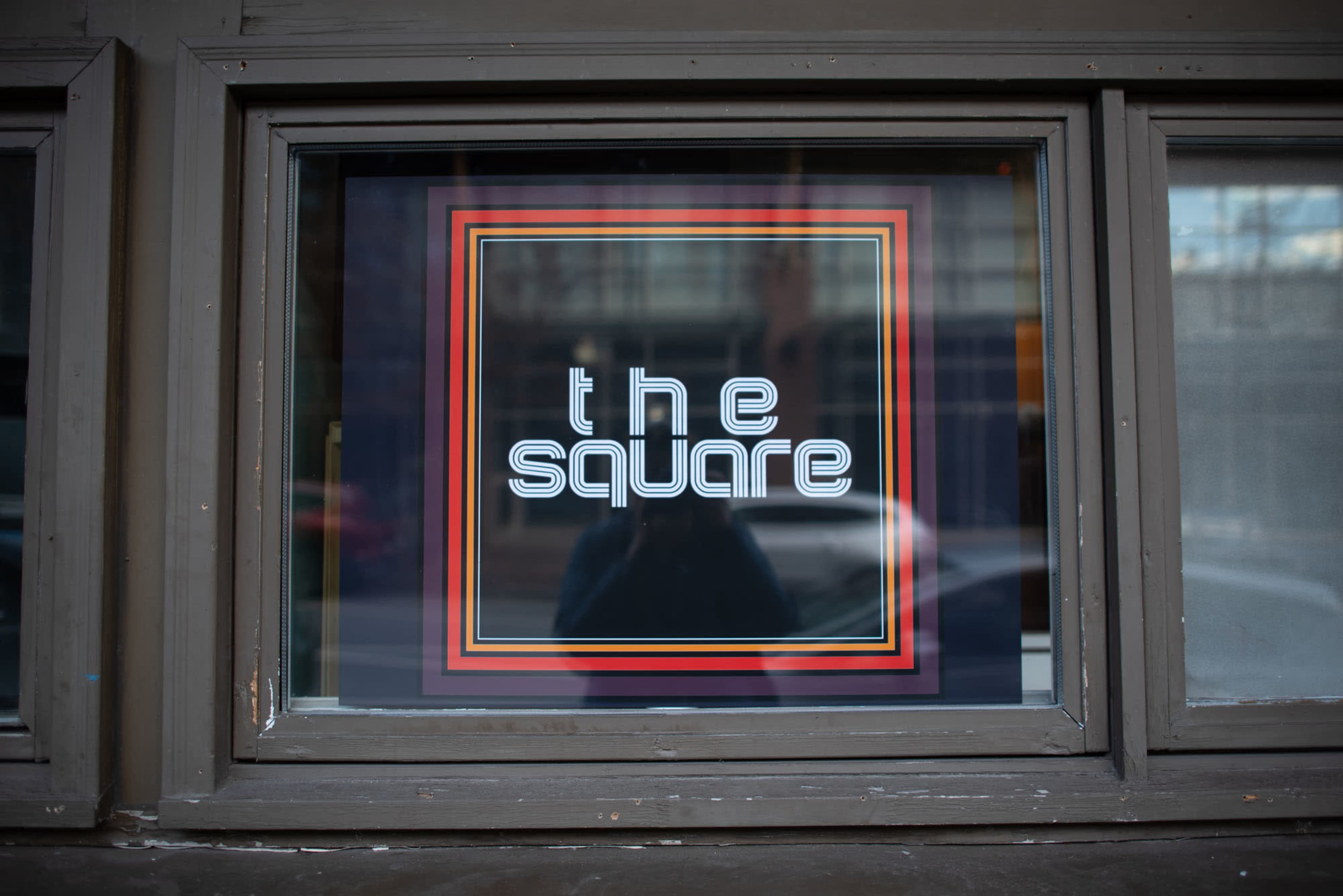 The image shows a window displaying a sign that reads "the square" in a bold, modern font with colorful borders.