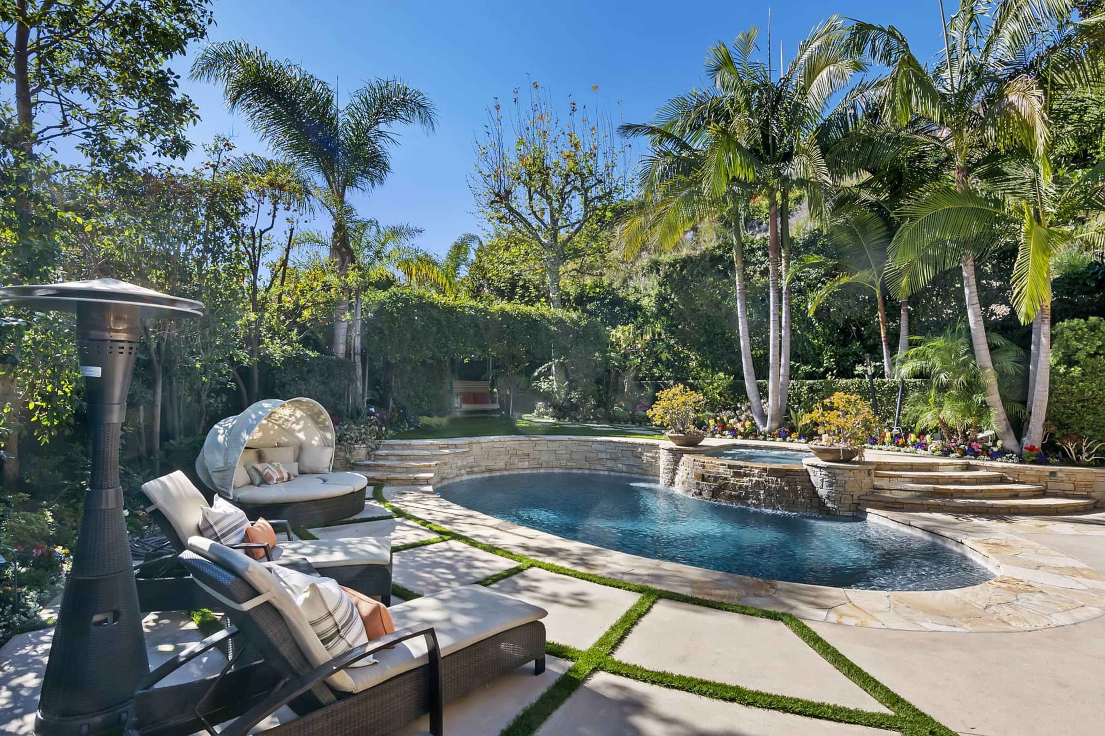 The image shows a landscaped backyard featuring a curved swimming pool, lounge chairs, a patio heater, and various tropical plants.