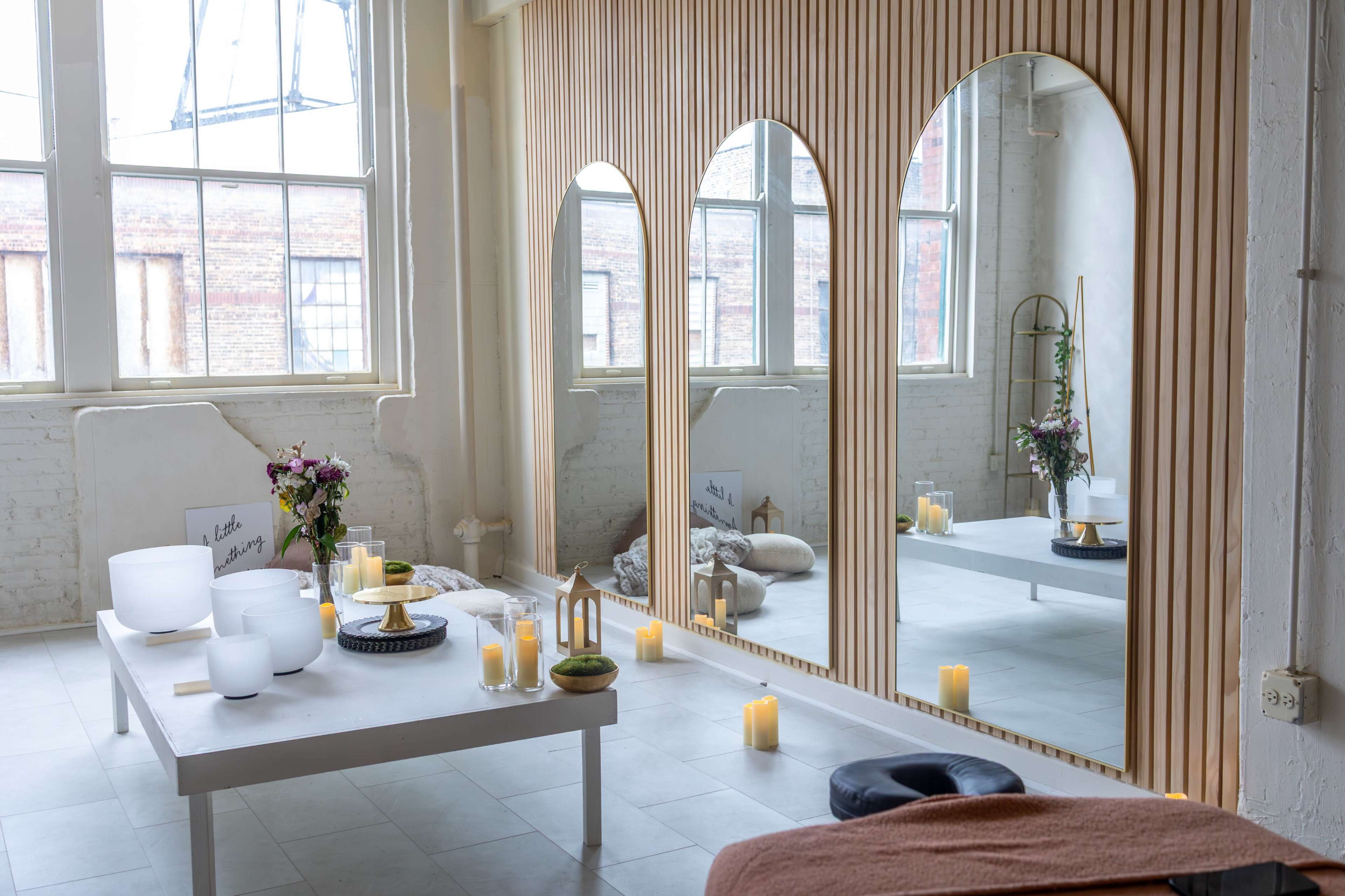 The image shows a serene indoor space featuring three large arched mirrors, a minimalist table with bowls, soft blankets, and candles arranged decoratively.