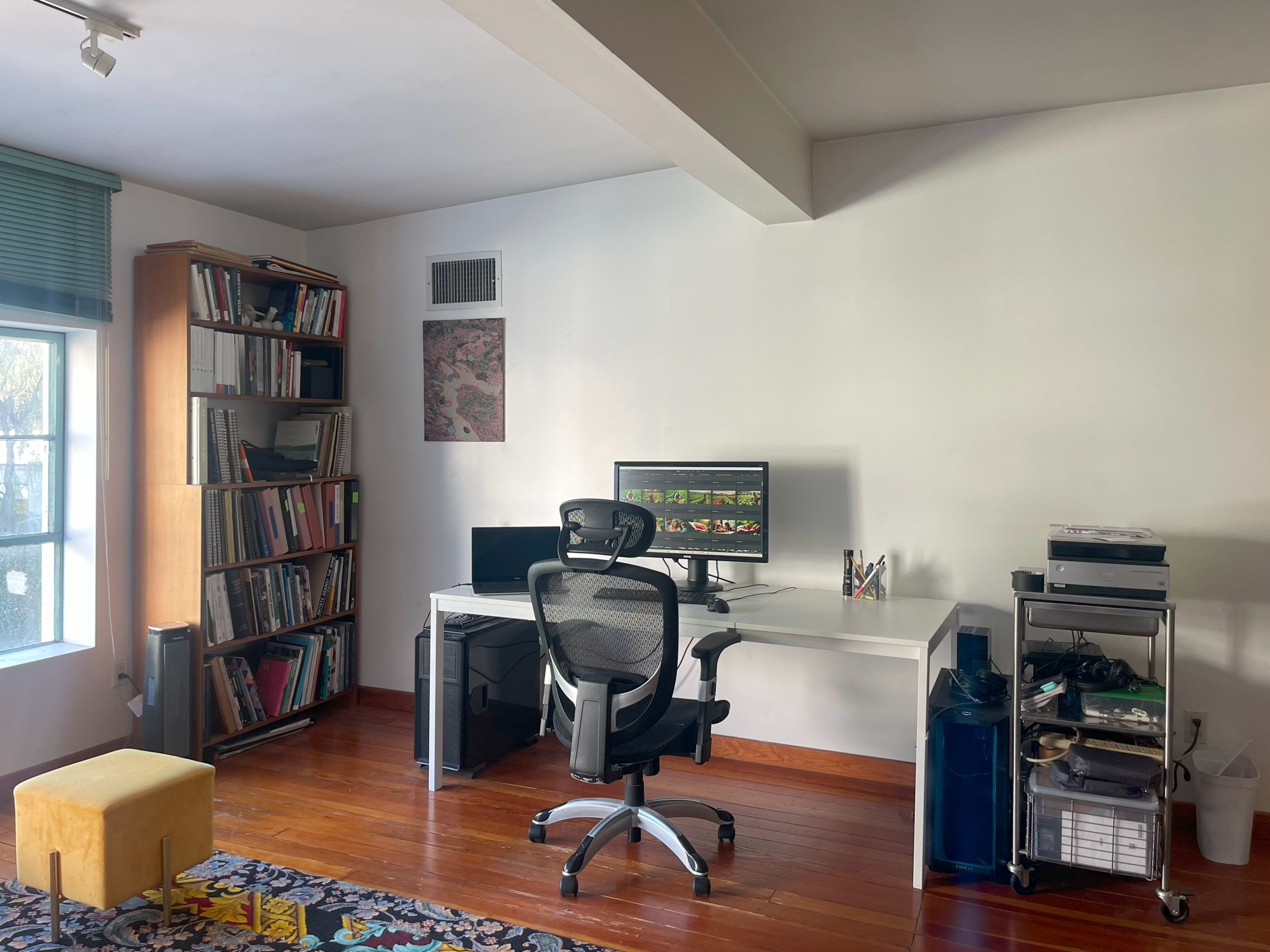 A modern office space featuring a desk with a computer, an ergonomic chair, a bookshelf filled with books, and a small yellow ottoman.