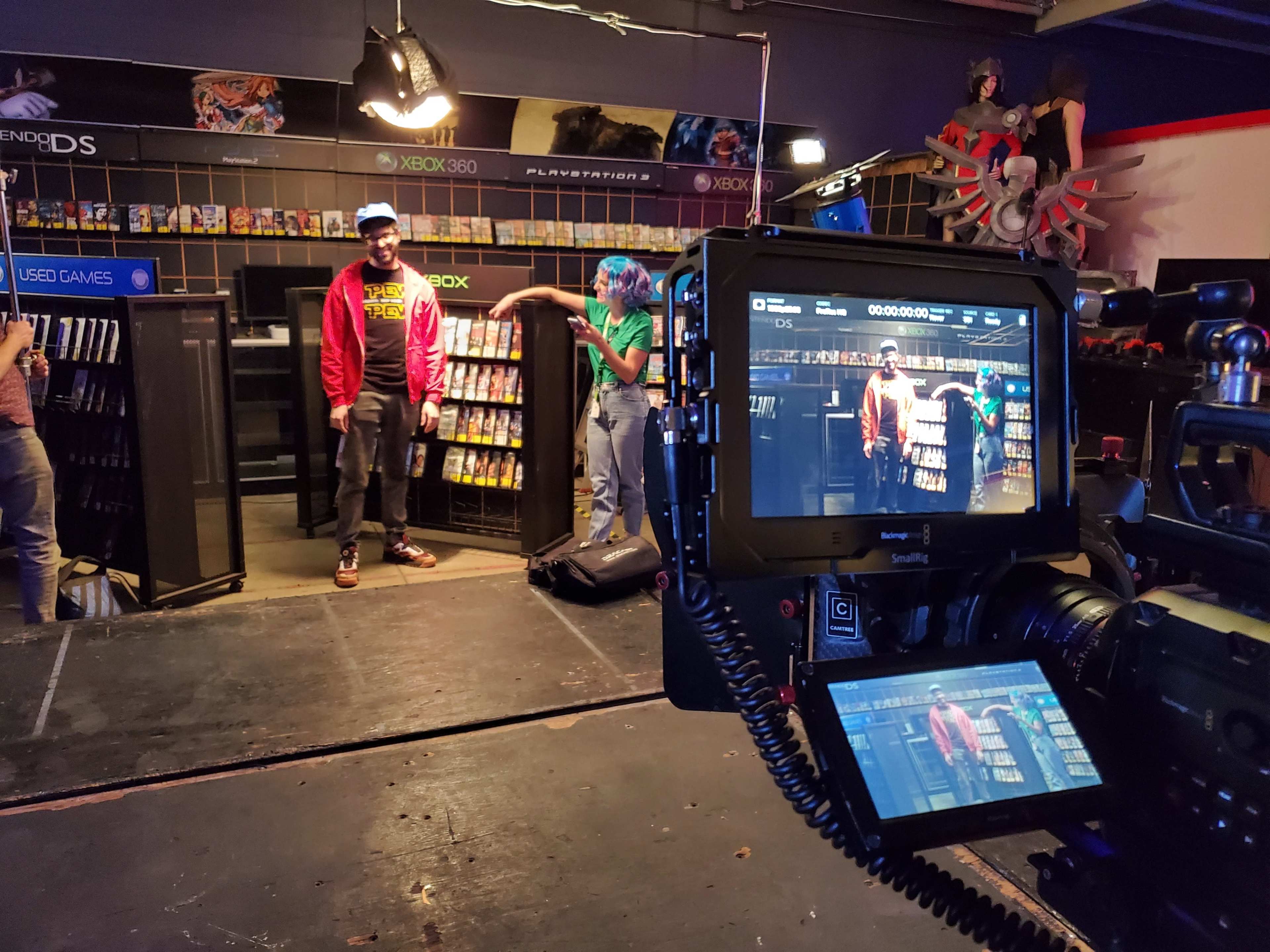 A film set featuring two people standing in a video game store aisle, with shelves filled with video games and a camera focused on them.