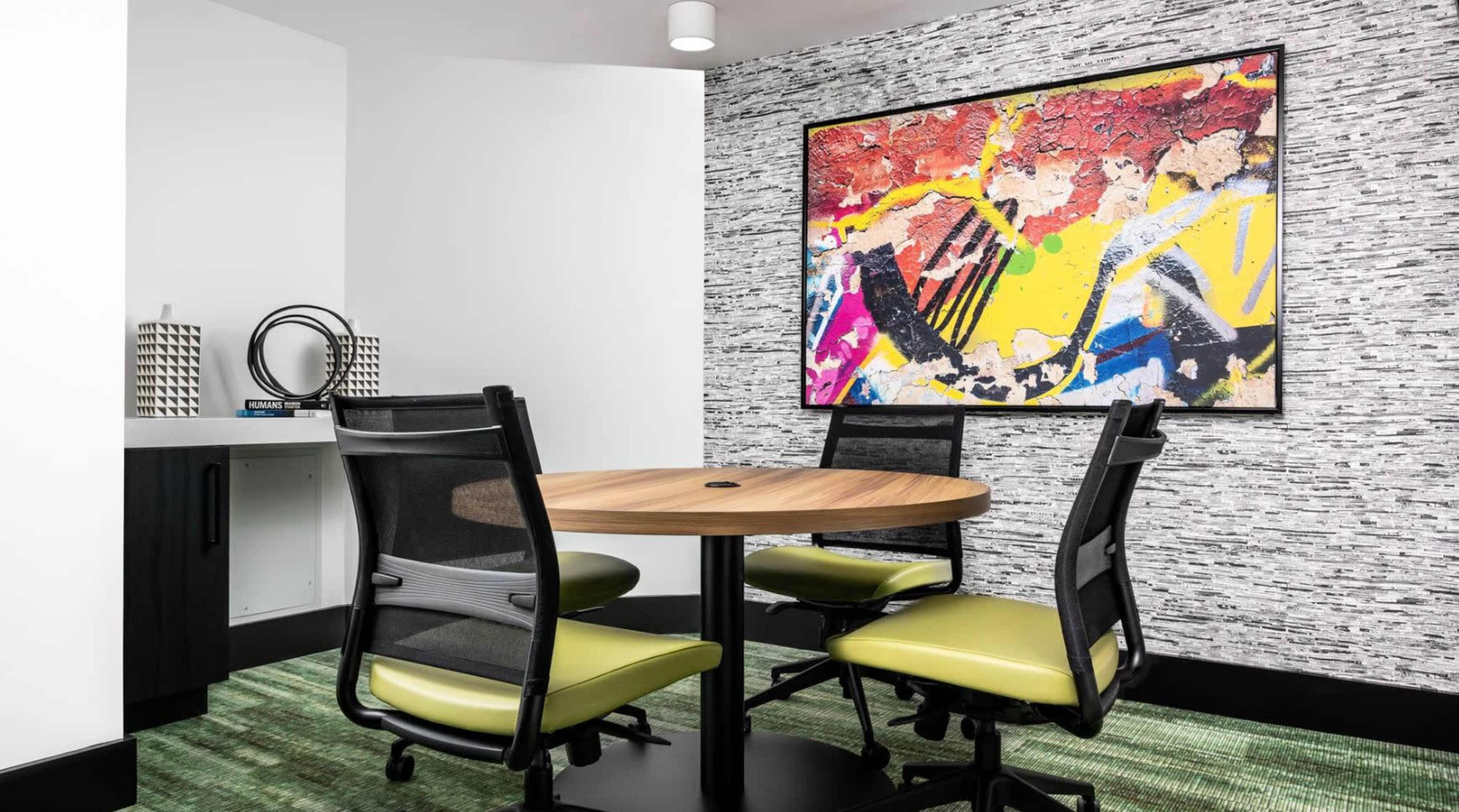 A conference room features a round wooden table surrounded by four black and green chairs, with a colorful abstract painting on the wall and textured wallpaper.