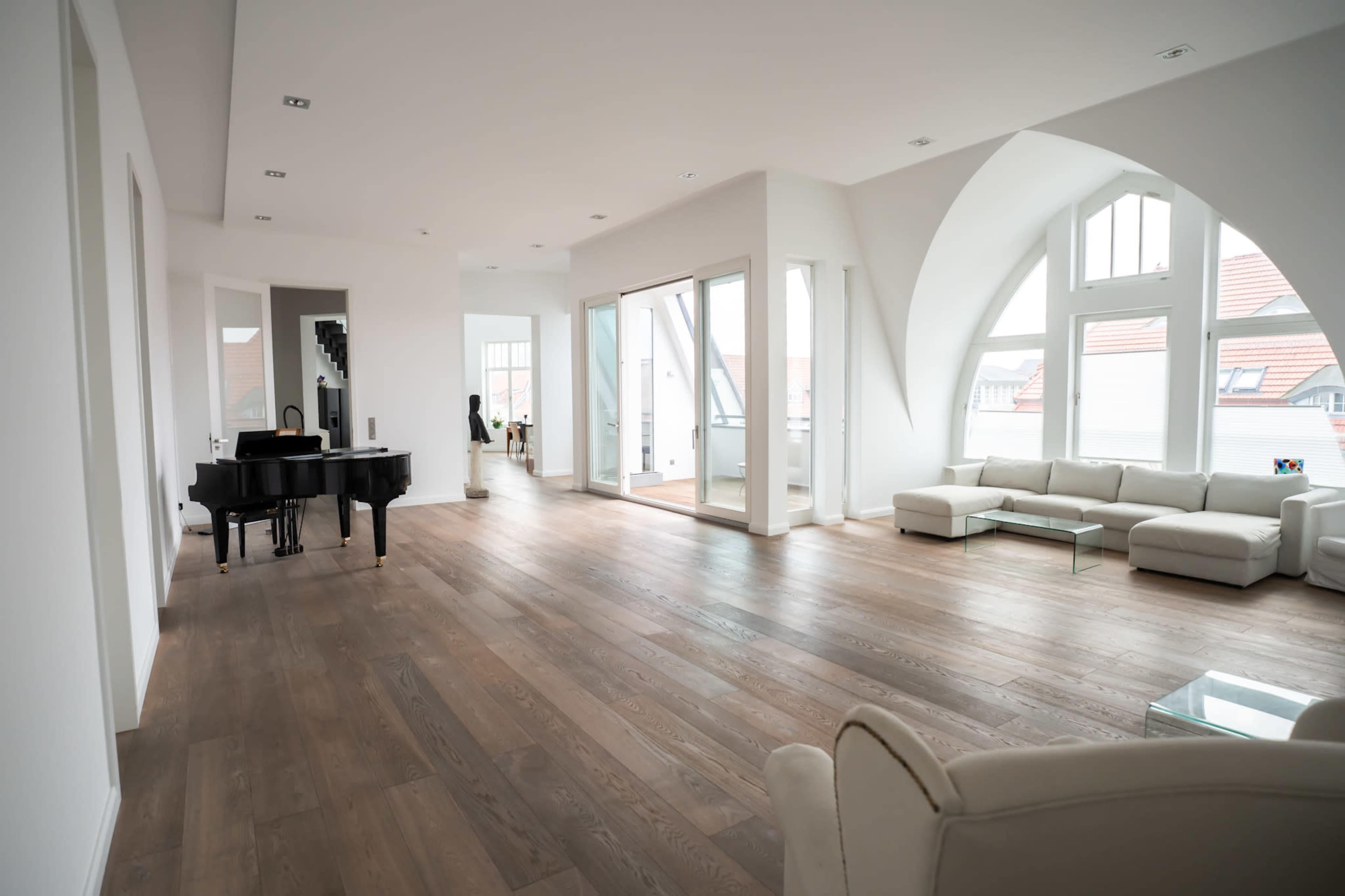 The image shows a spacious, modern living area with a grand piano, light-colored furniture, and large windows allowing ample natural light.