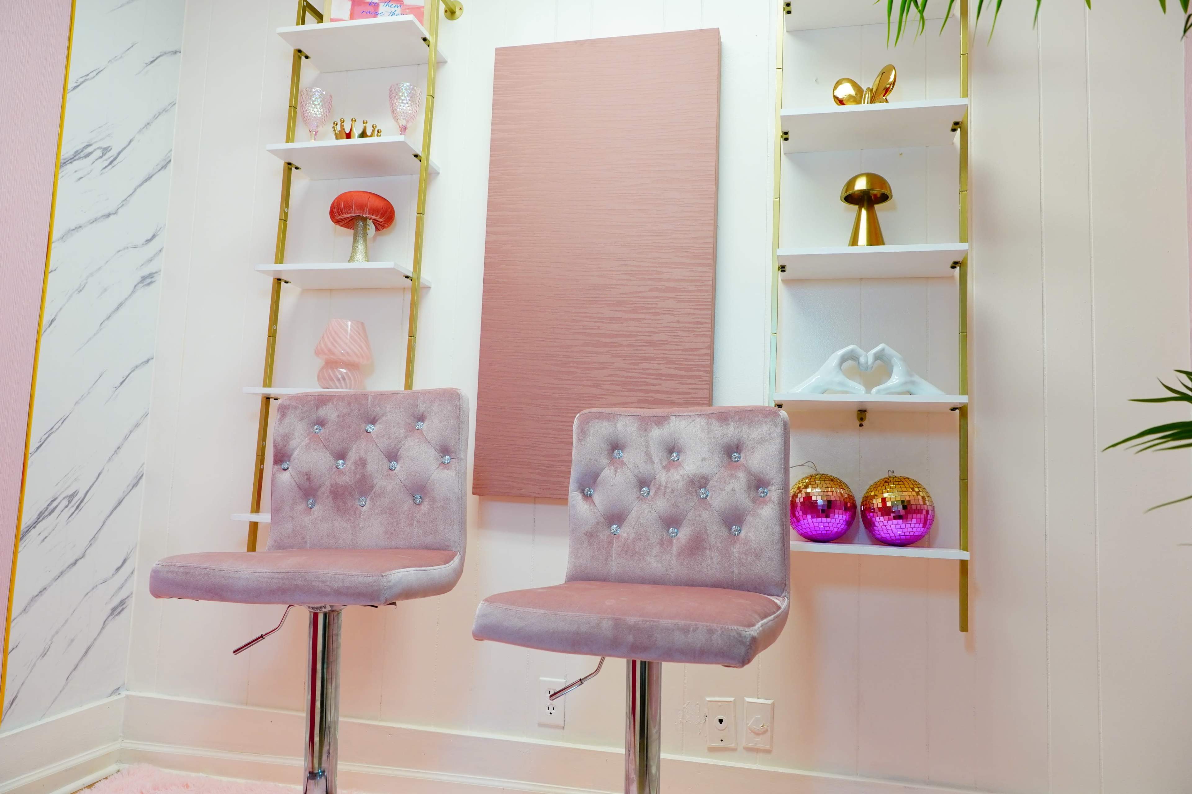 Two plush barstools sit in front of a minimalist shelf display with various decorative items against a light-colored wall.