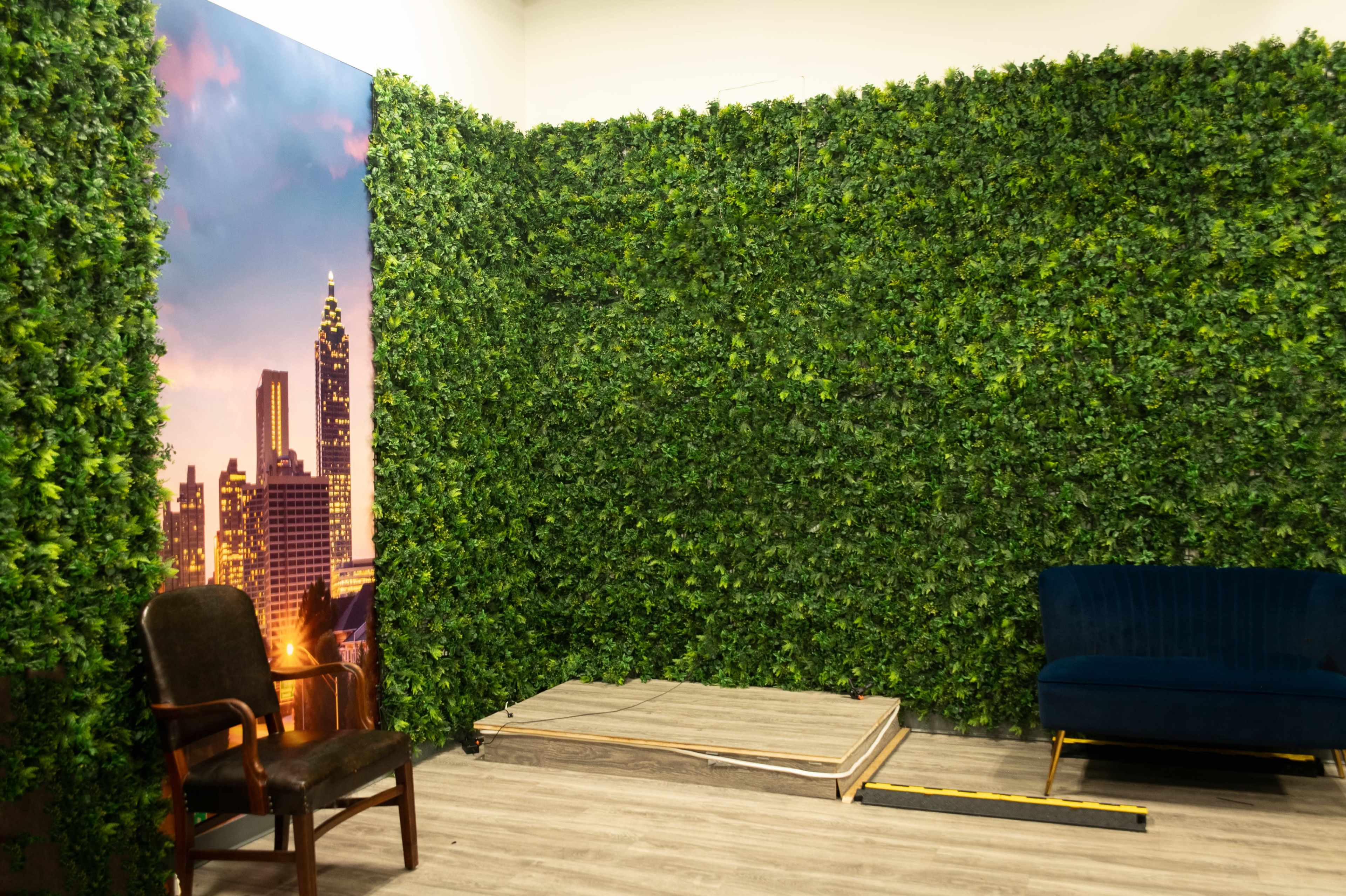 The image shows a room with a wall covered in artificial greenery, featuring a skyline backdrop and a sofa next to a wooden platform.