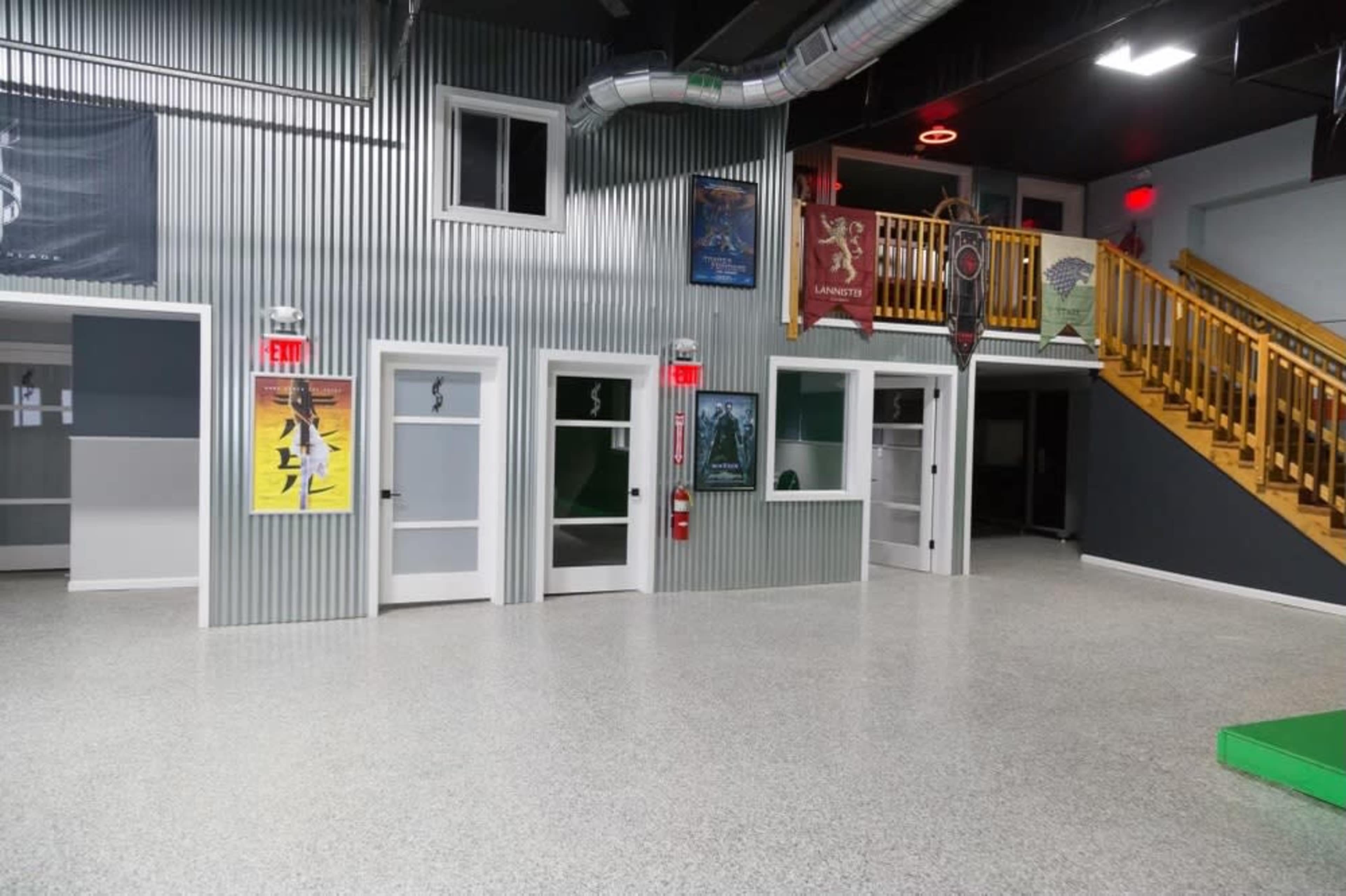 A spacious indoor area with metal-paneled walls, several closed doors, and posters displayed on the walls, along with a staircase leading to an upper level.