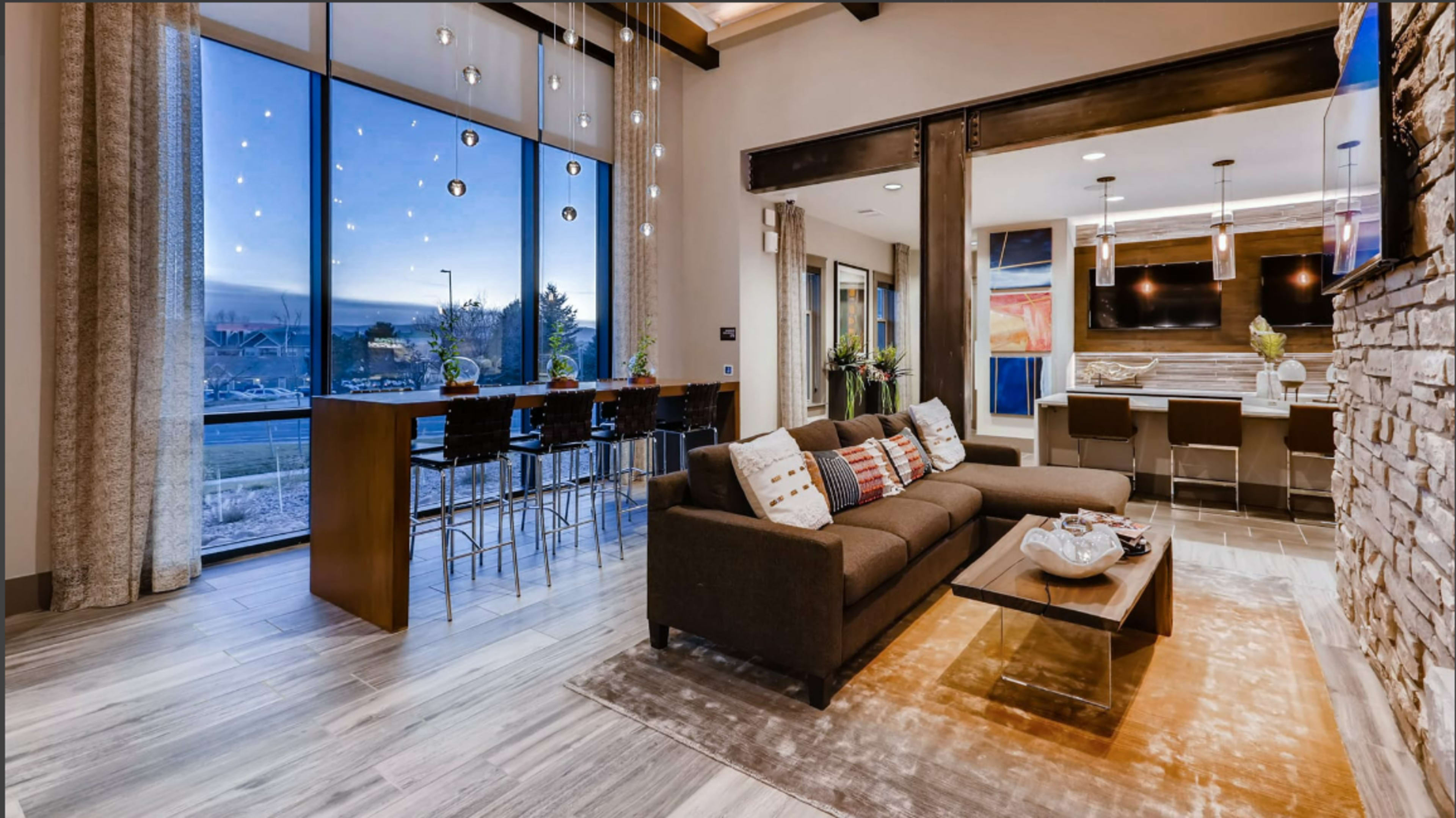 A stylish living area featuring a brown sofa, a coffee table, and a dining space with bar stools, all illuminated by pendant lights and large windows offering a view of the outdoors.