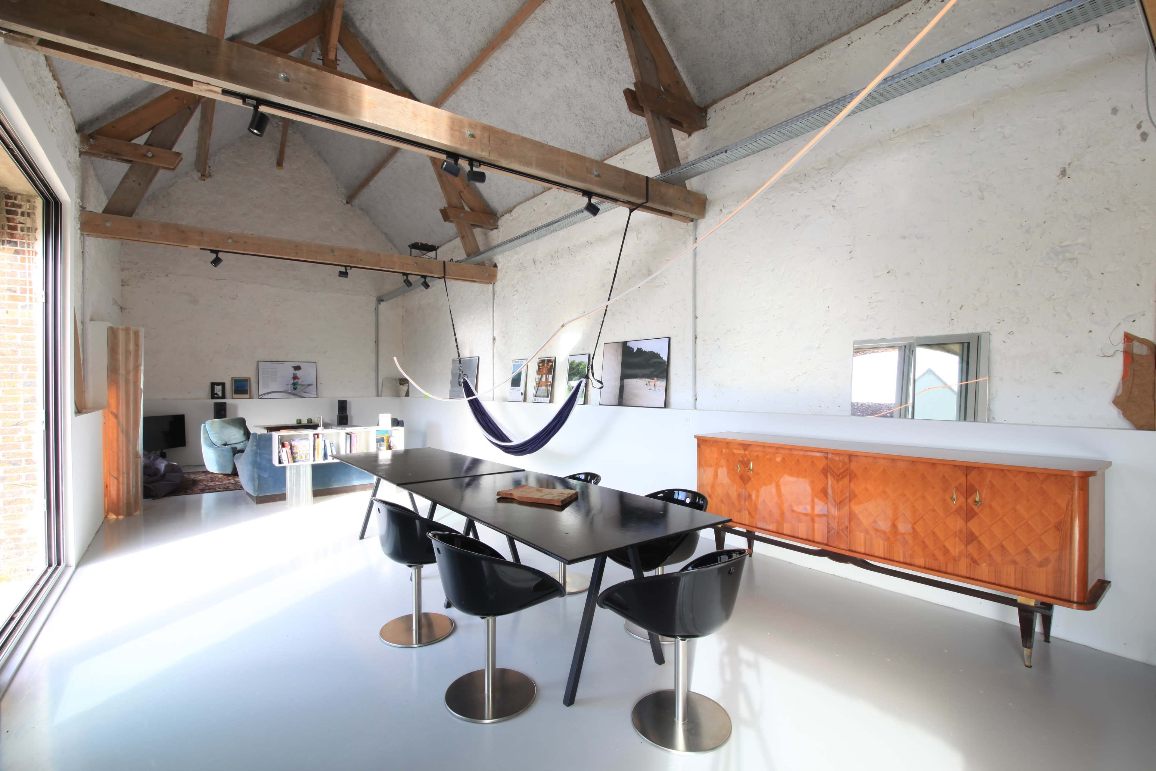 A spacious, modern interior with a high ceiling featuring wooden beams, a dining table surrounded by black chairs, a hammock, and a wooden sideboard against the wall.