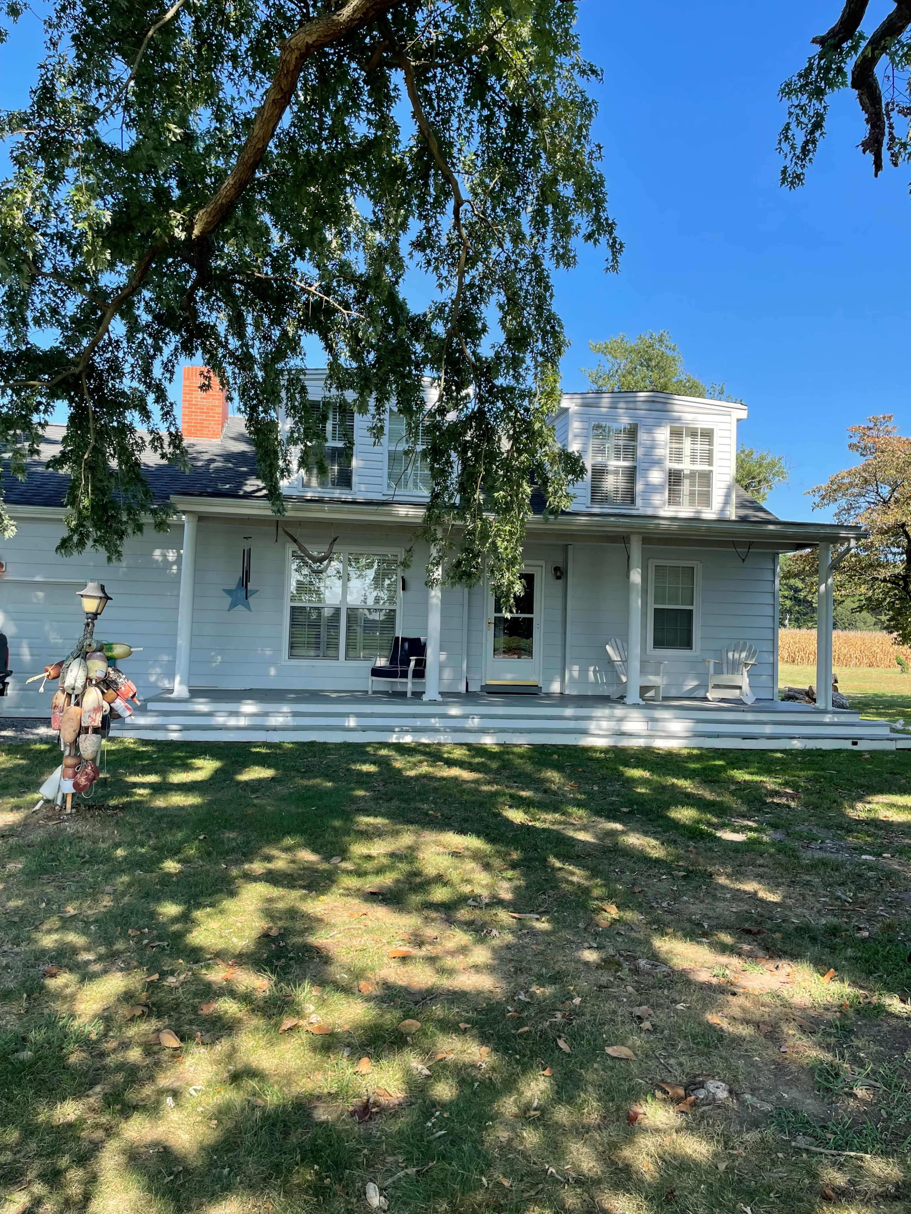A two-story house with a covered porch and large windows is situated under a tree, showcasing a green lawn and a field in the background.