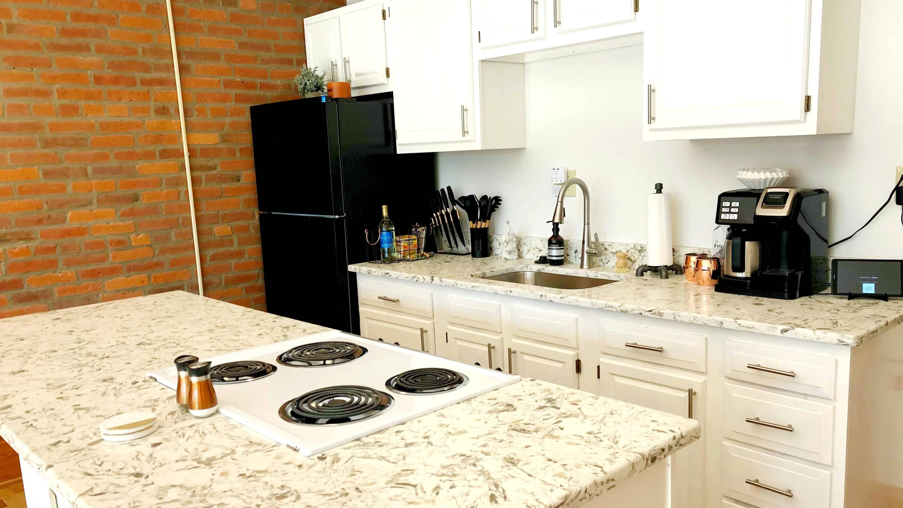 The kitchen features a dark refrigerator, white cabinets, a granite countertop with a stove, and various kitchen appliances arranged on the countertops.