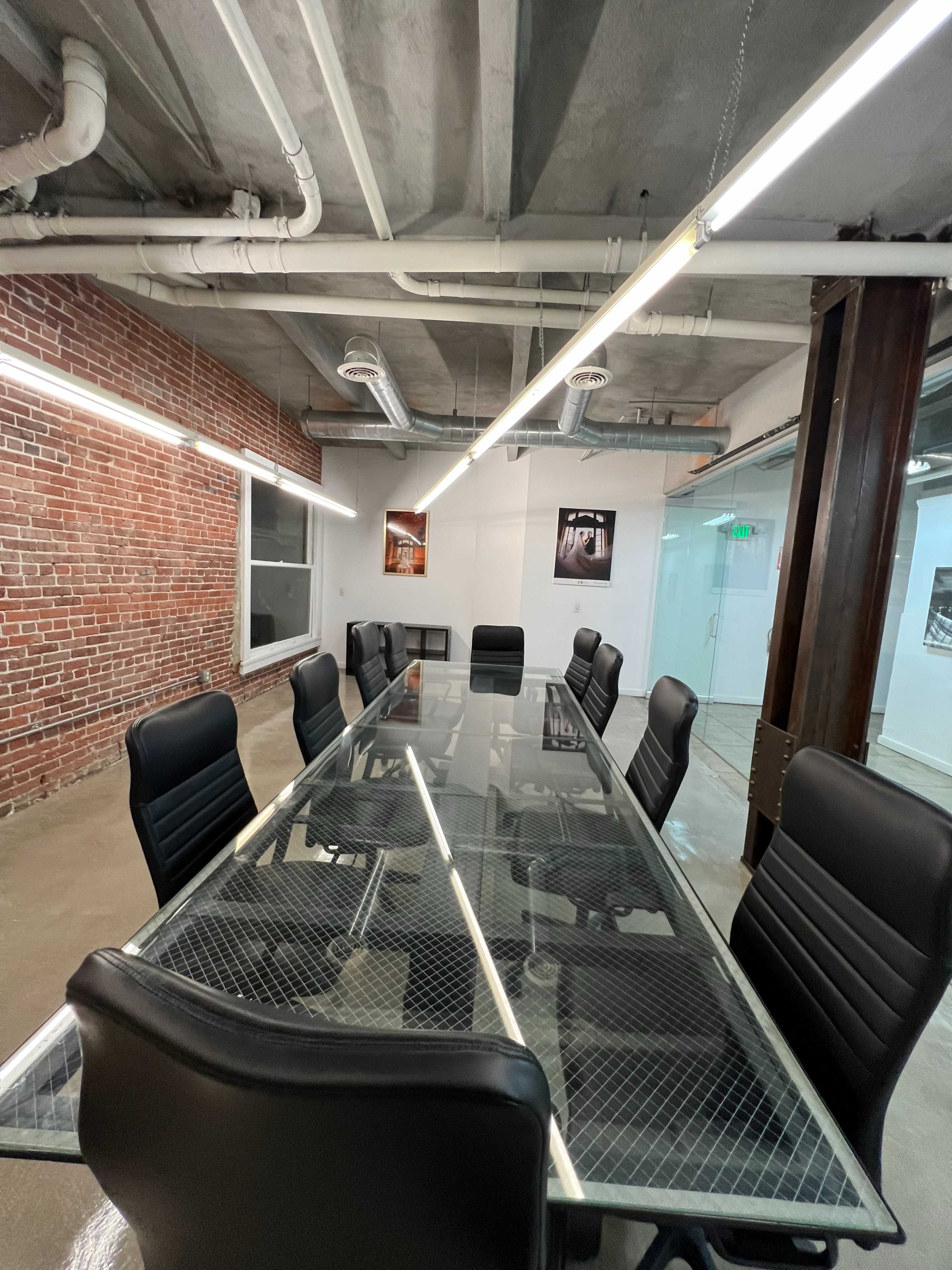 A modern conference room features a long glass table surrounded by black ergonomic chairs, with exposed brick walls and overhead lighting.