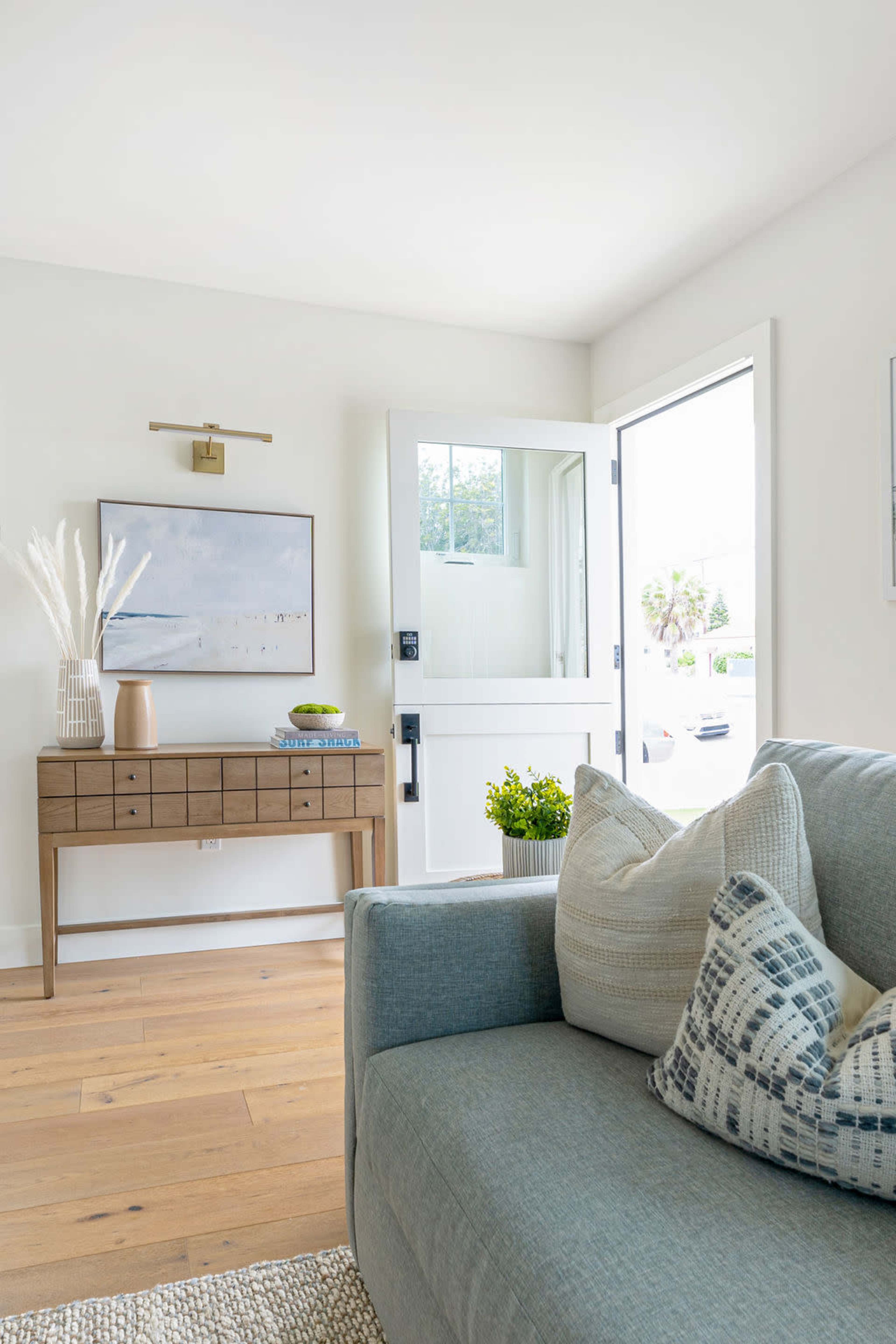 A bright living room features a light-colored sofa with decorative pillows, a wooden console table against the wall, and an open door leading to the outside.