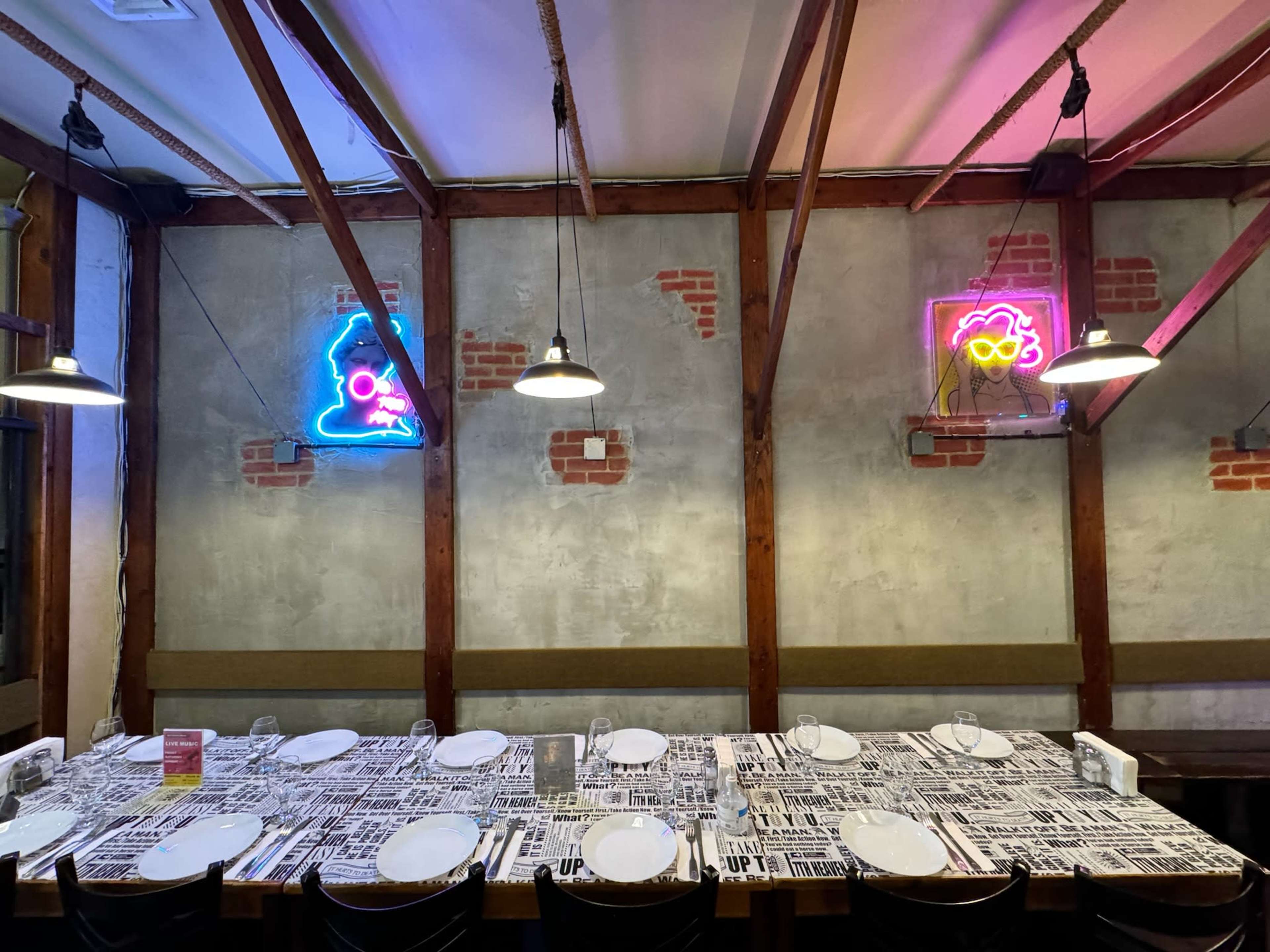 The image shows a restaurant interior with exposed brick walls, wooden beams, and neon signs of a figure and glasses, illuminated above a table set with cutlery and menus.