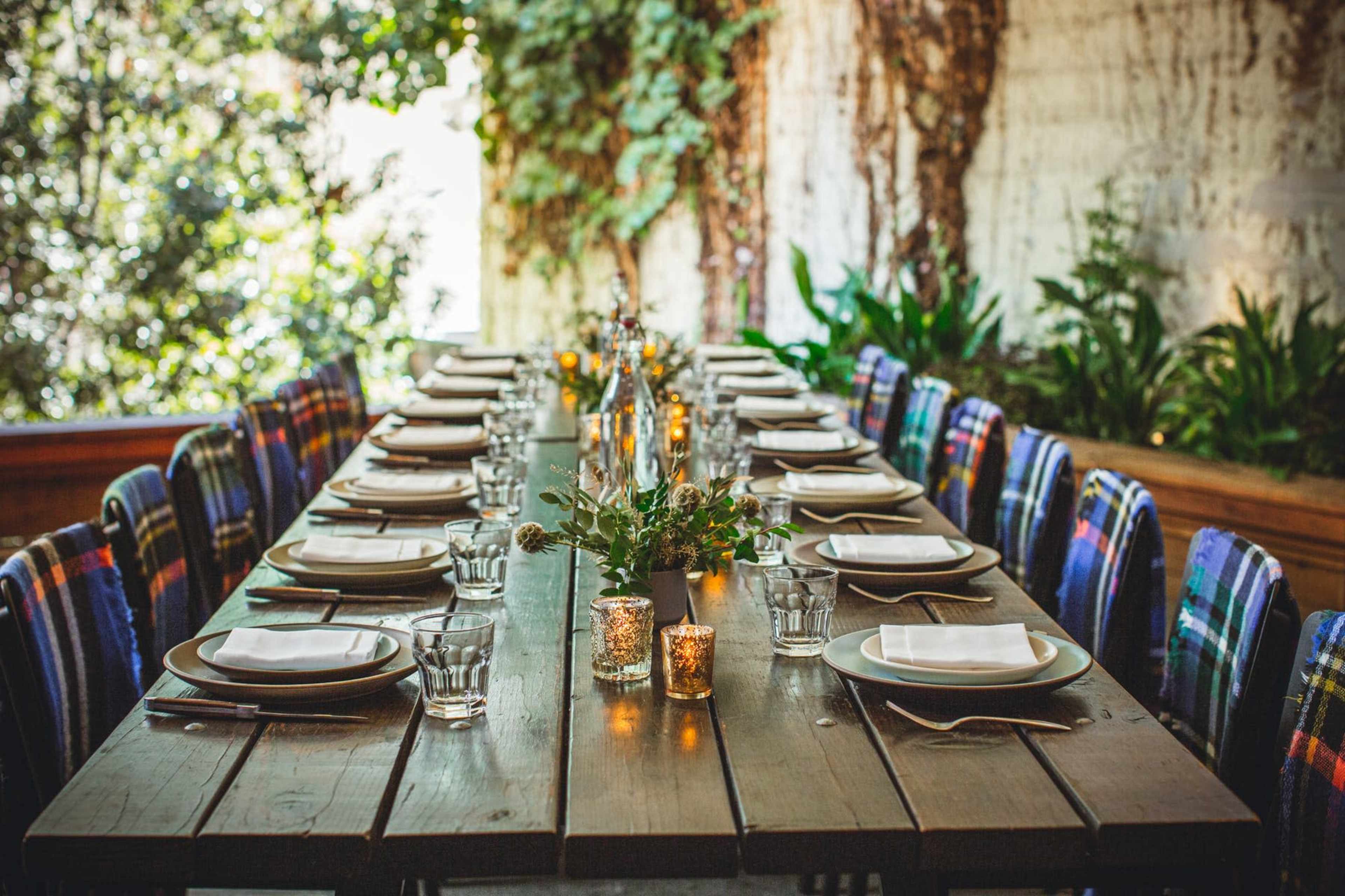 A long wooden dining table is set with plates and glasses, adorned with small candleholders and centerpieces, surrounded by colorful chairs in a lush, green environment.