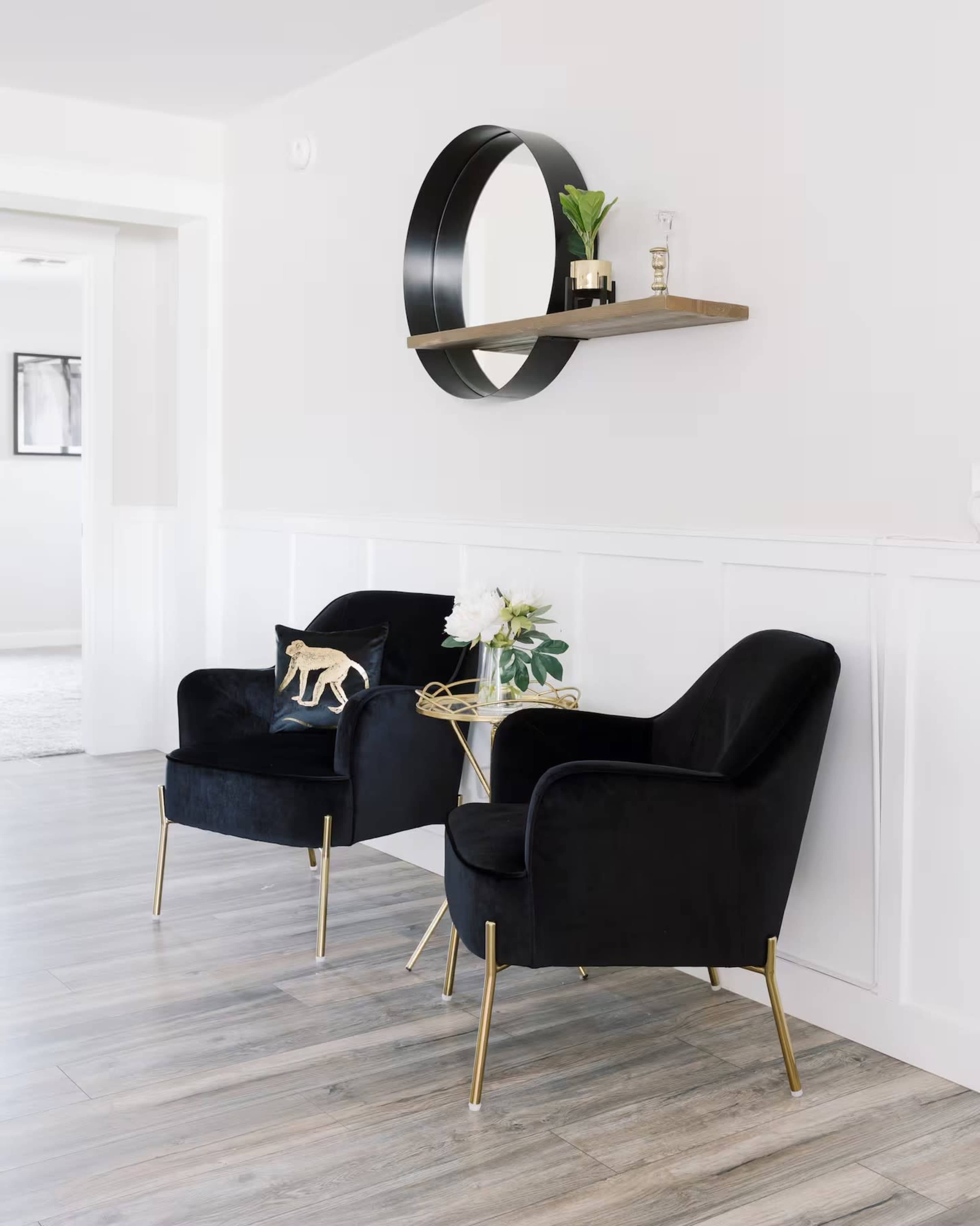 The image features two black velvet chairs with gold legs situated beside a small round table, under a wall-mounted shelf with a circular mirror above it.