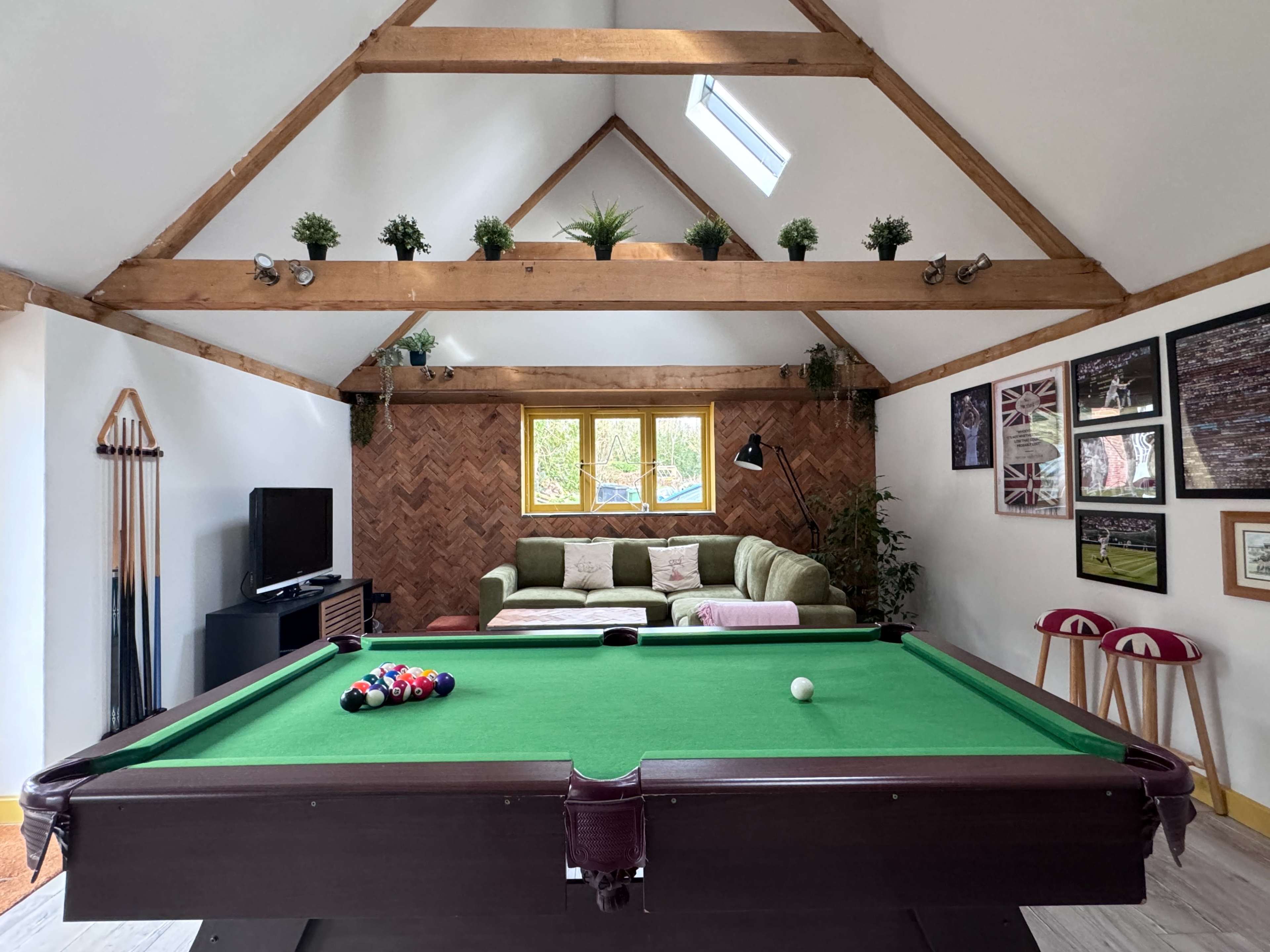 A games room with a green pool table in the foreground, a brown herringbone wall, and a seating area with a green sofa and plants against a white wall.