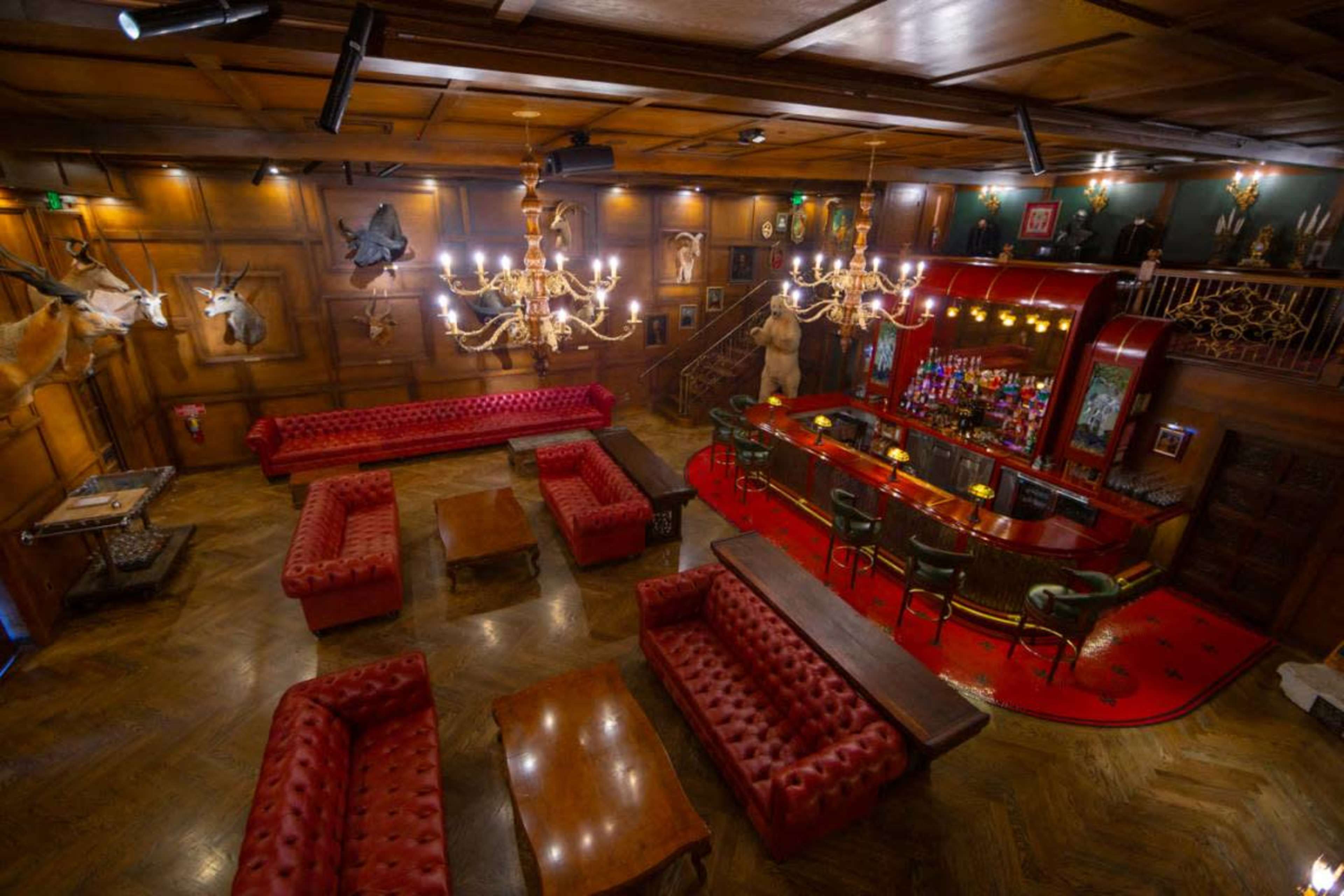 The image depicts an elegant lounge with red tufted sofas, a wooden bar, and mounted animal heads on the walls.