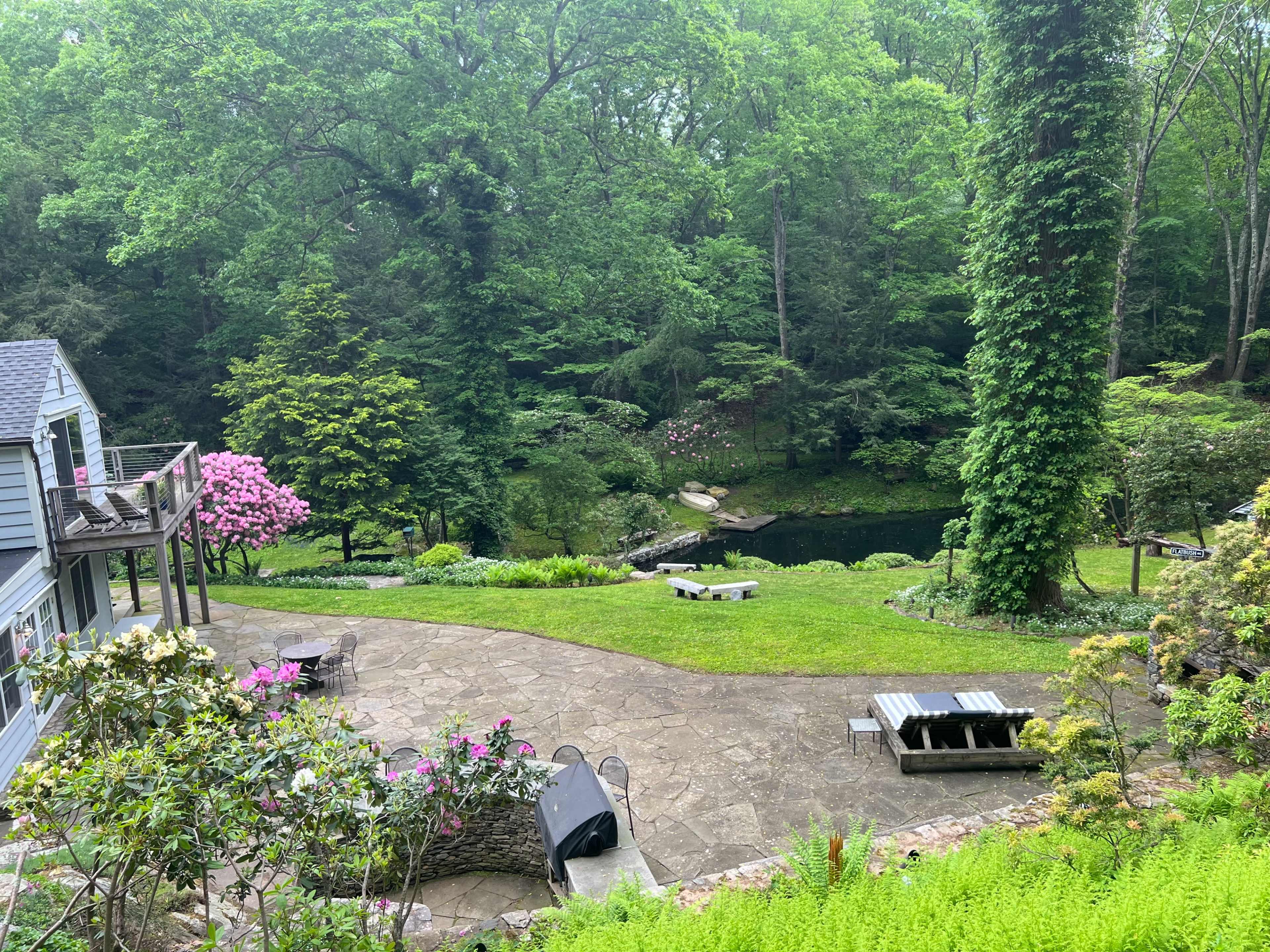 A landscaped backyard with stone pathways, seating areas, flowering plants, and a pond surrounded by trees.