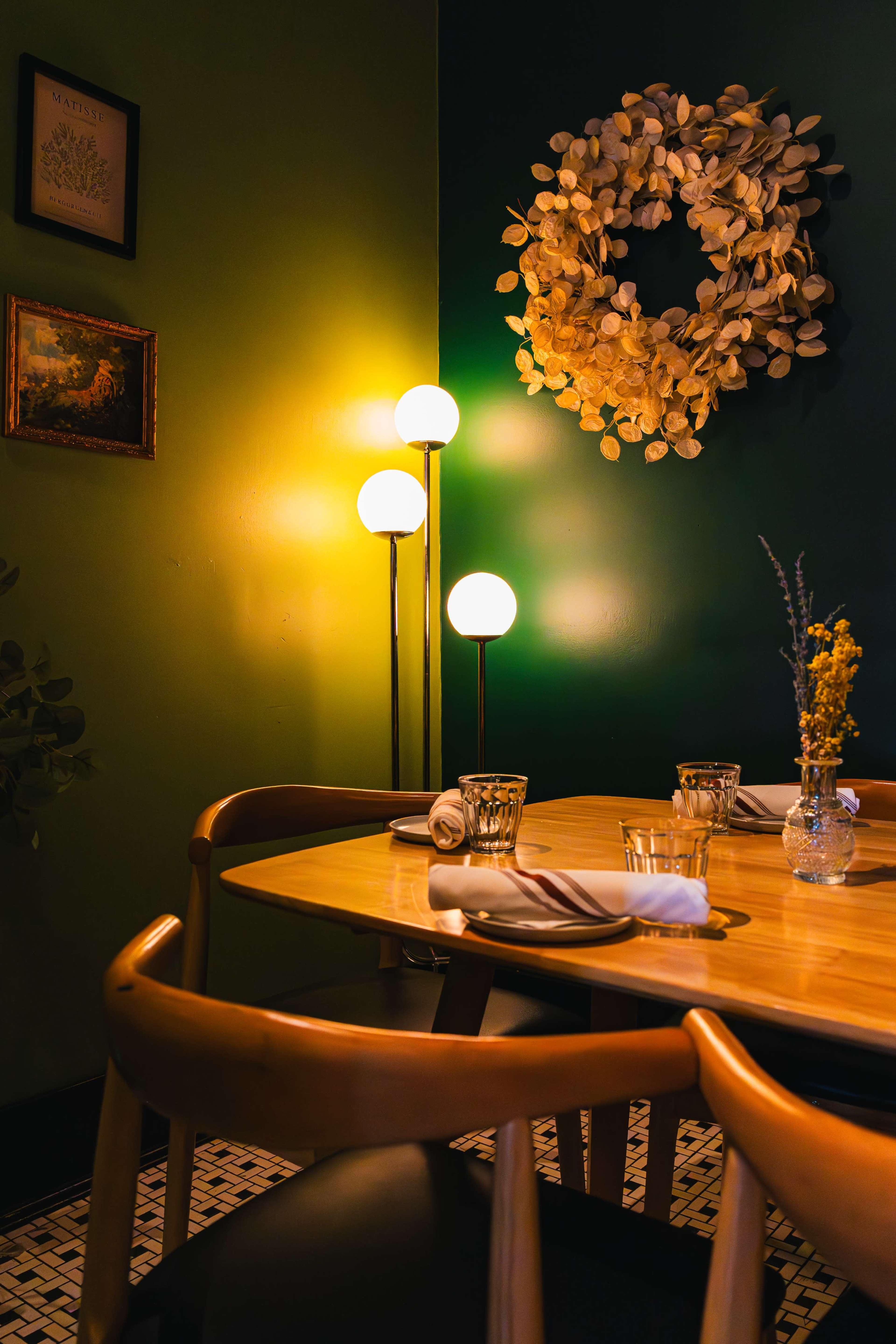 A wooden dining table is set with glassware and utensils, illuminated by a standing lamp and a wall-mounted wreath in a cozy, green-walled restaurant.