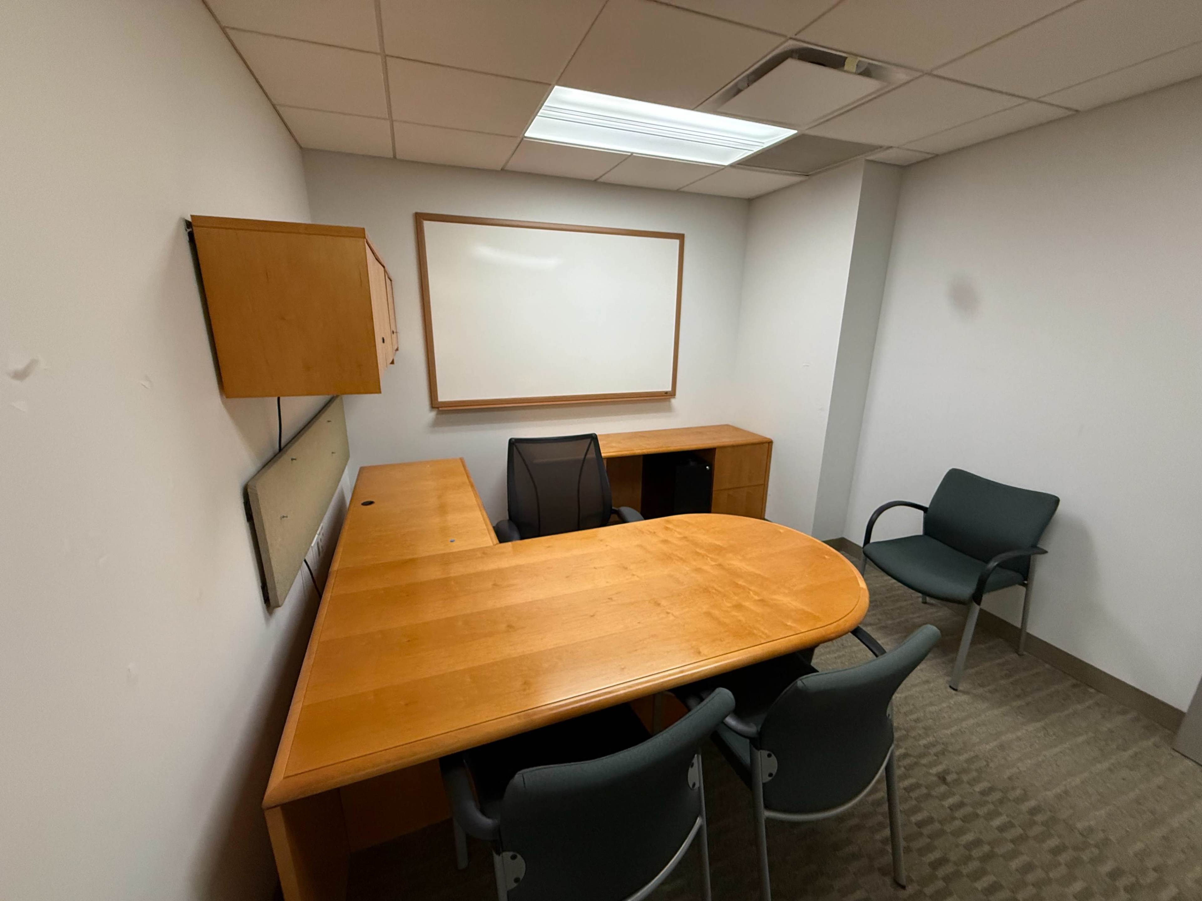 The image shows a small, empty meeting room with a wooden desk, a chair, and a whiteboard on the wall.