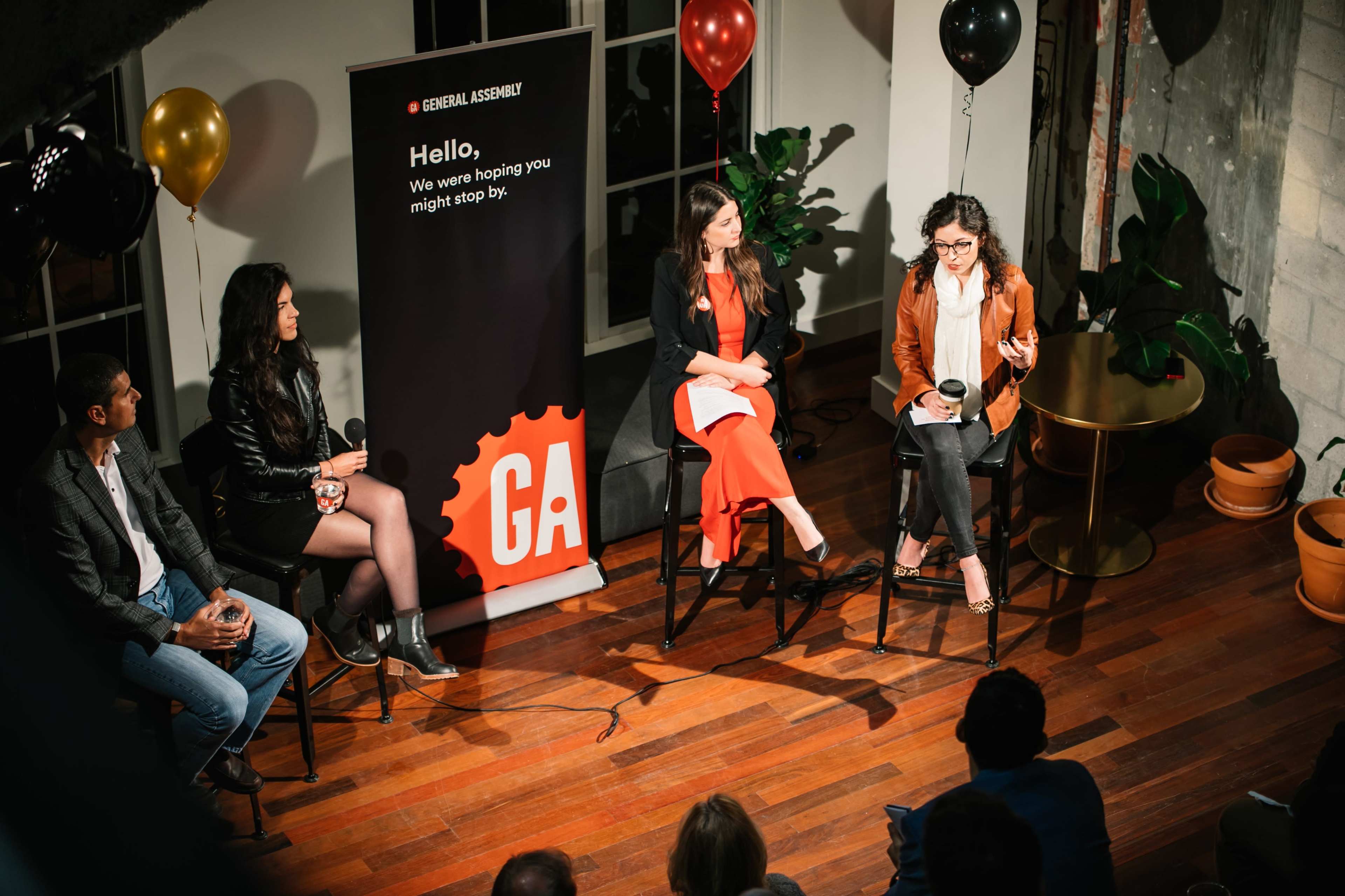 A panel discussion takes place in a venue featuring four speakers seated on chairs, with a microphone and a backdrop promoting General Assembly.