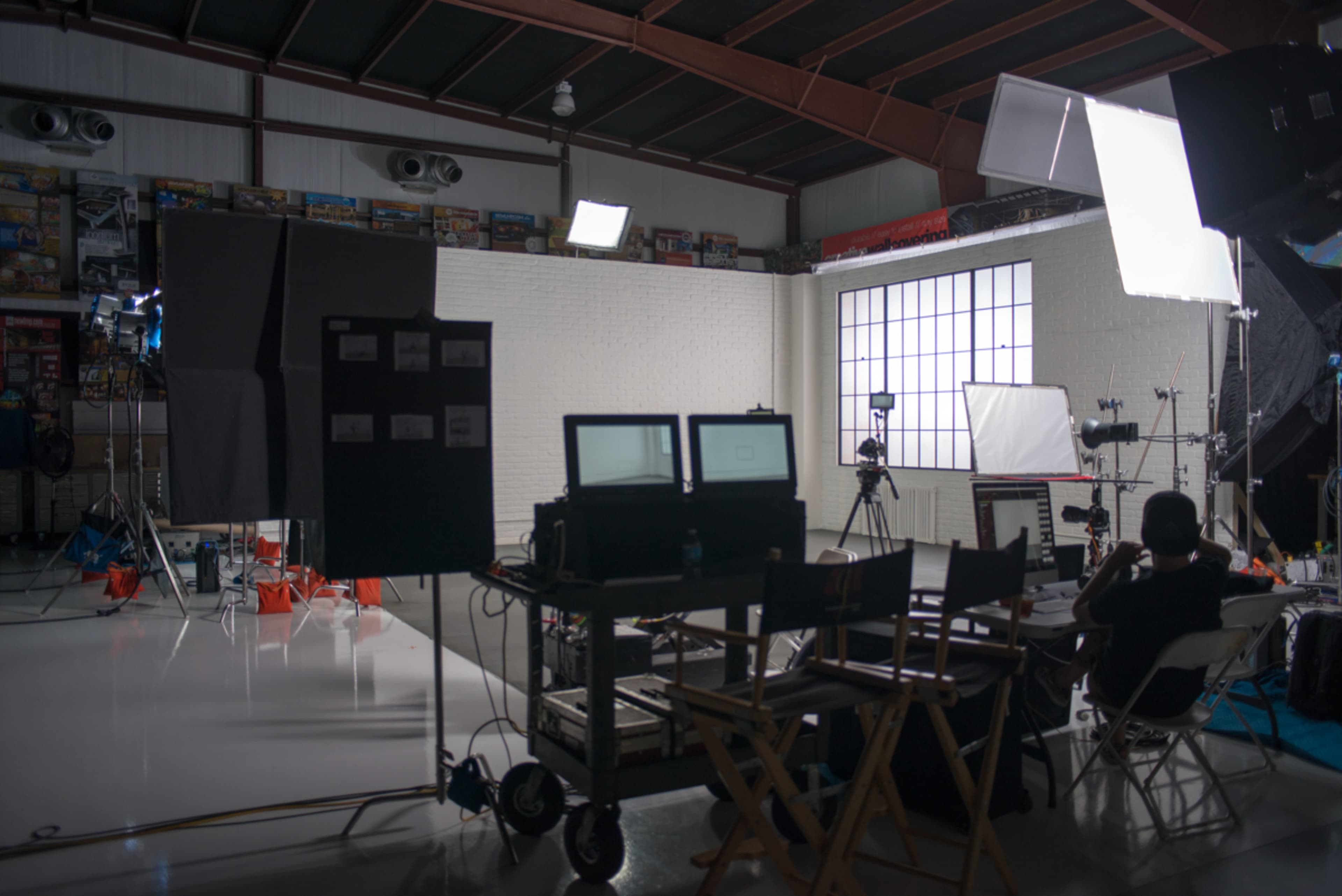 A film set features multiple cameras, lighting equipment, and two monitors arranged on a cart in a spacious studio with a large window.