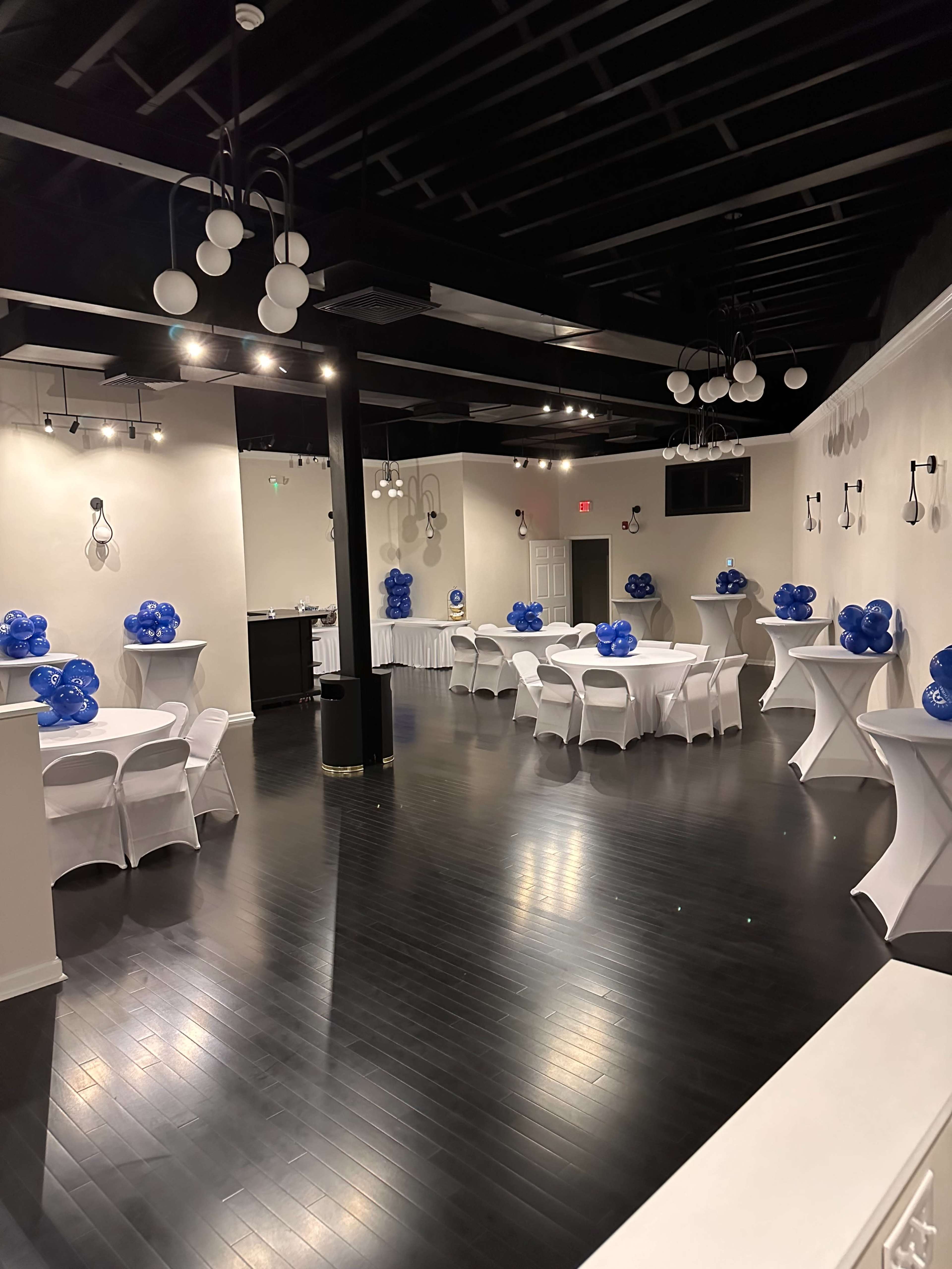 The image shows a spacious event venue with white tables adorned with blue balloons, set against a black wooden floor and illuminated by ceiling lights.