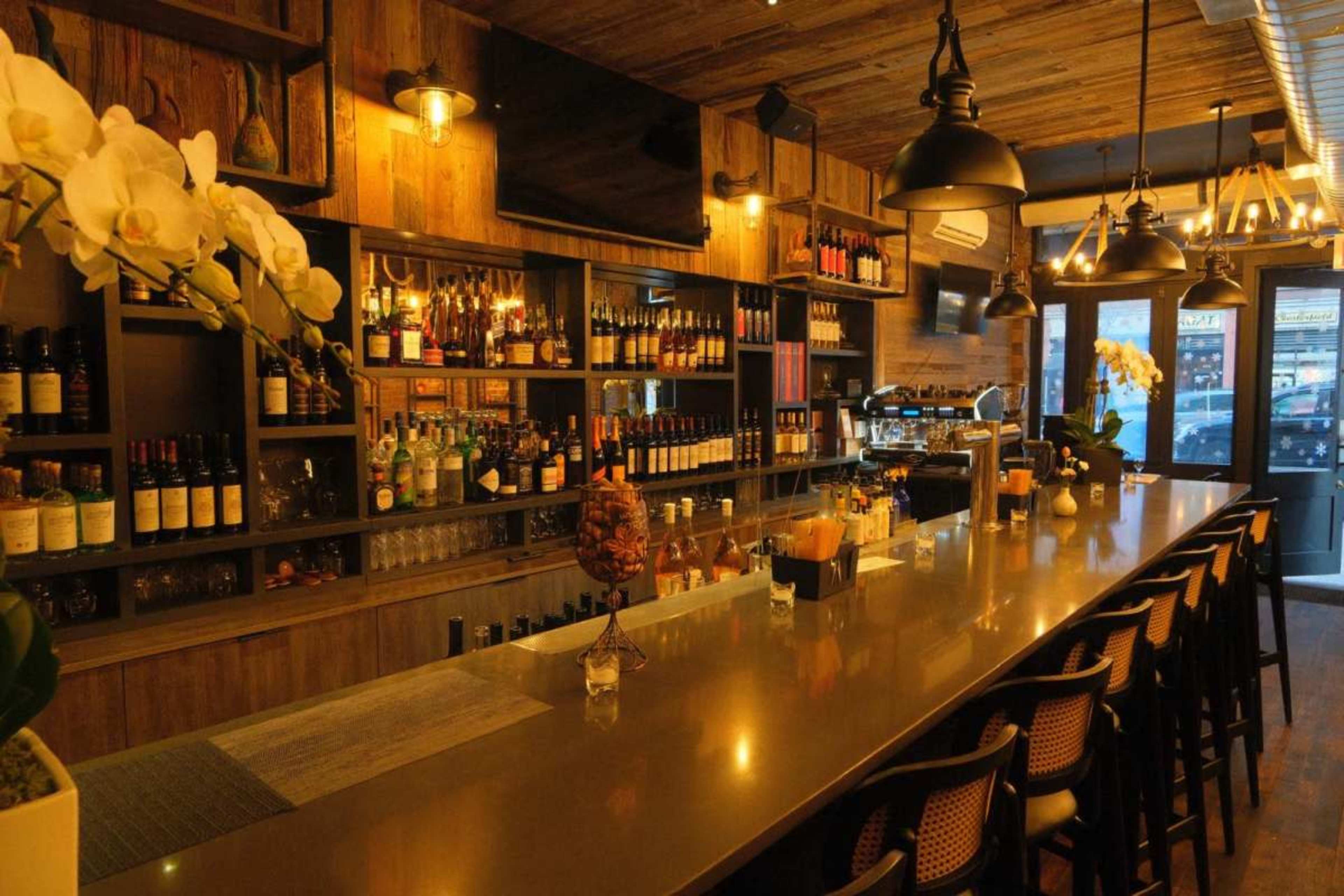 The image shows a stylish bar with a long, polished countertop, shelves filled with various bottles of liquor, and warm wood-paneled walls.