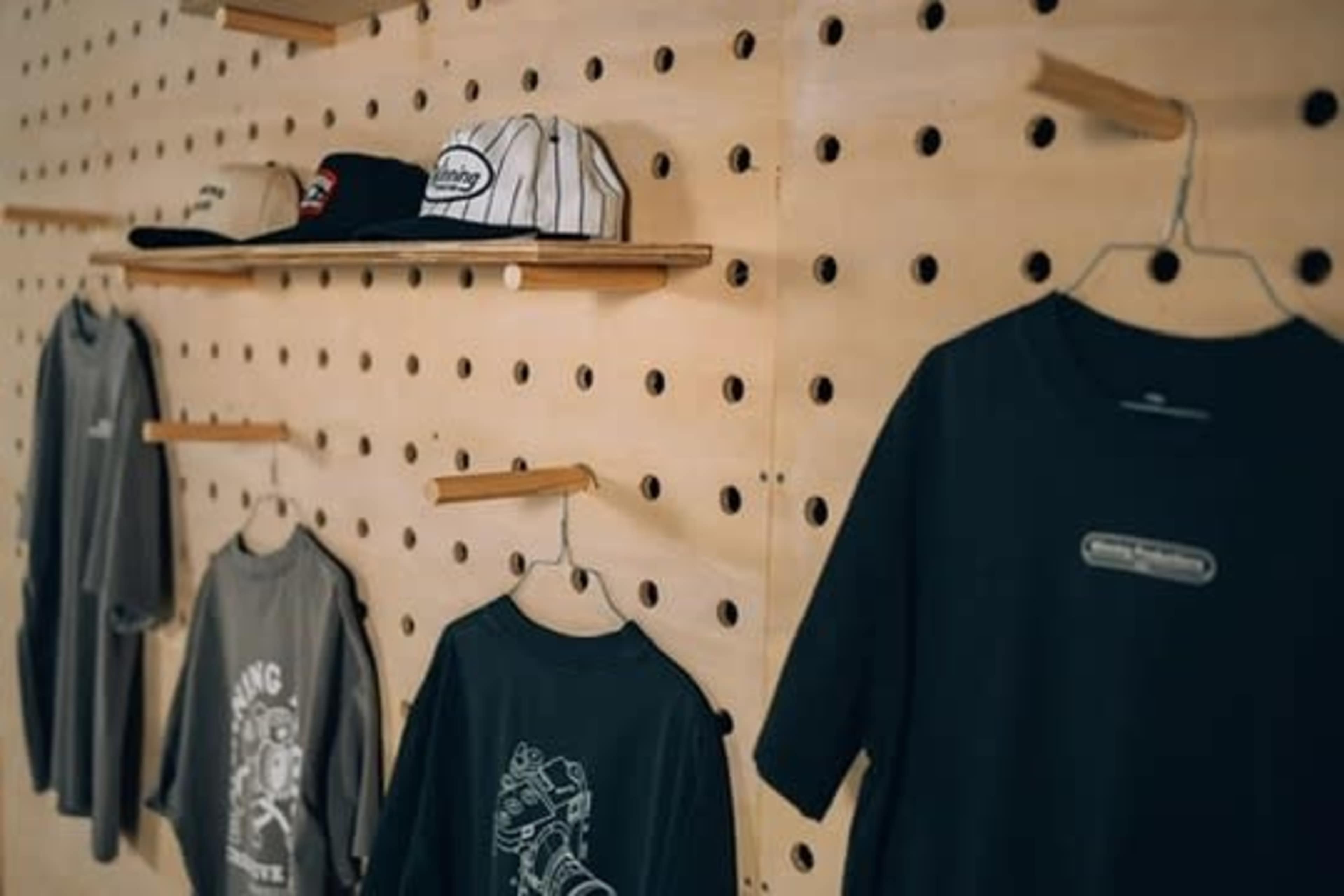 The image shows a wooden wall with a series of holes displaying hats on shelves and T-shirts hanging below.