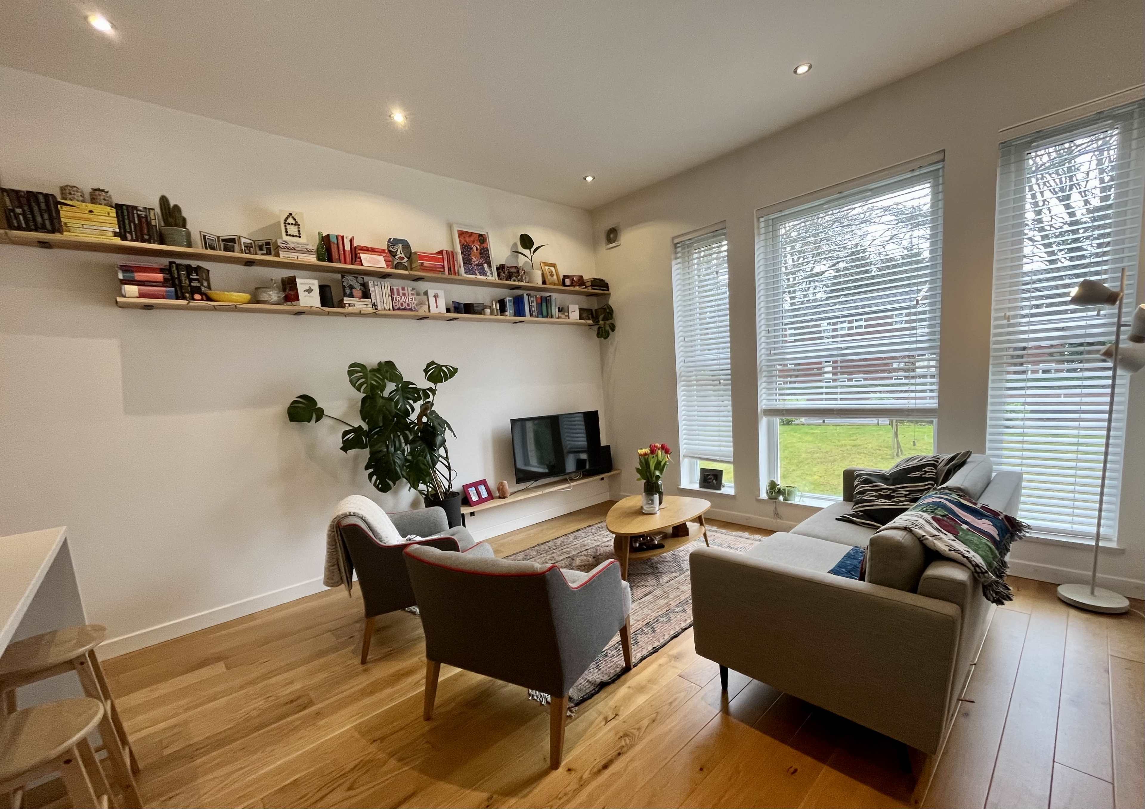 A cozy living room features a gray sofa, two armchairs, a wooden coffee table, and a long shelf adorned with books and decor.