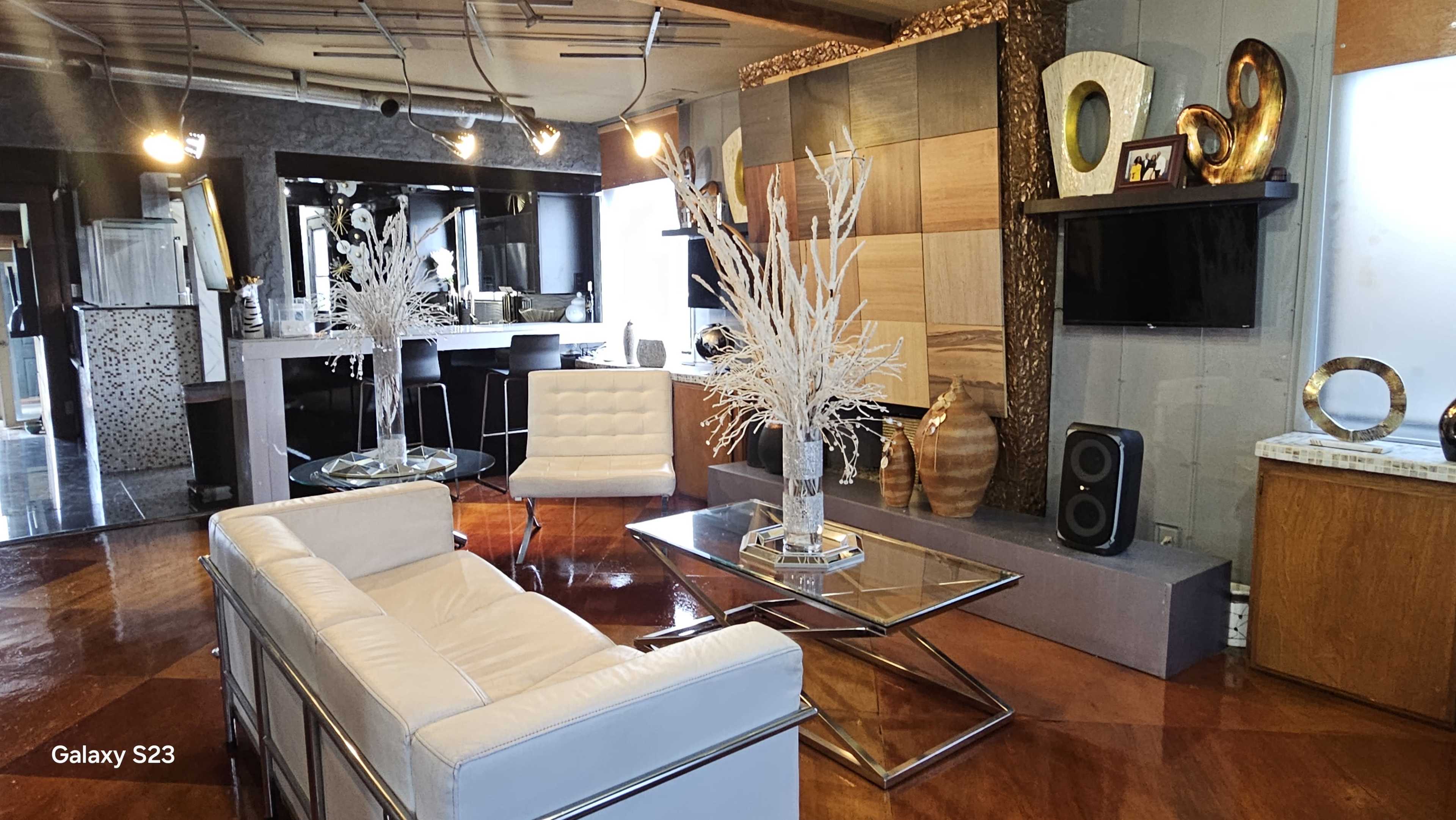 The image shows a modern living room featuring a white leather sofa, glass coffee table, decorative elements, and a flat-screen TV mounted on the wall.