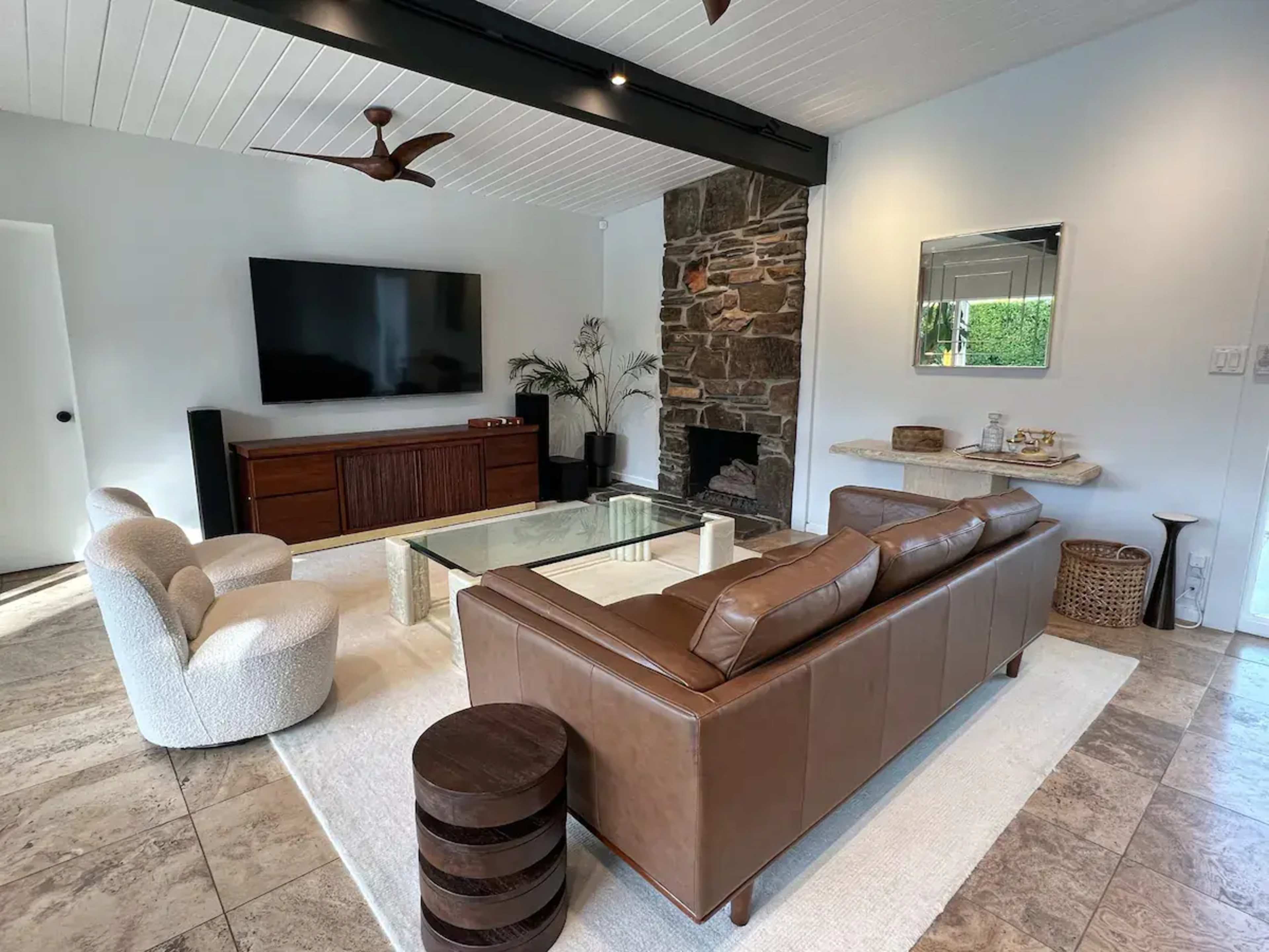 The image shows a modern living room featuring a brown leather sofa, a glass coffee table, a stone fireplace, and a wall-mounted television.
