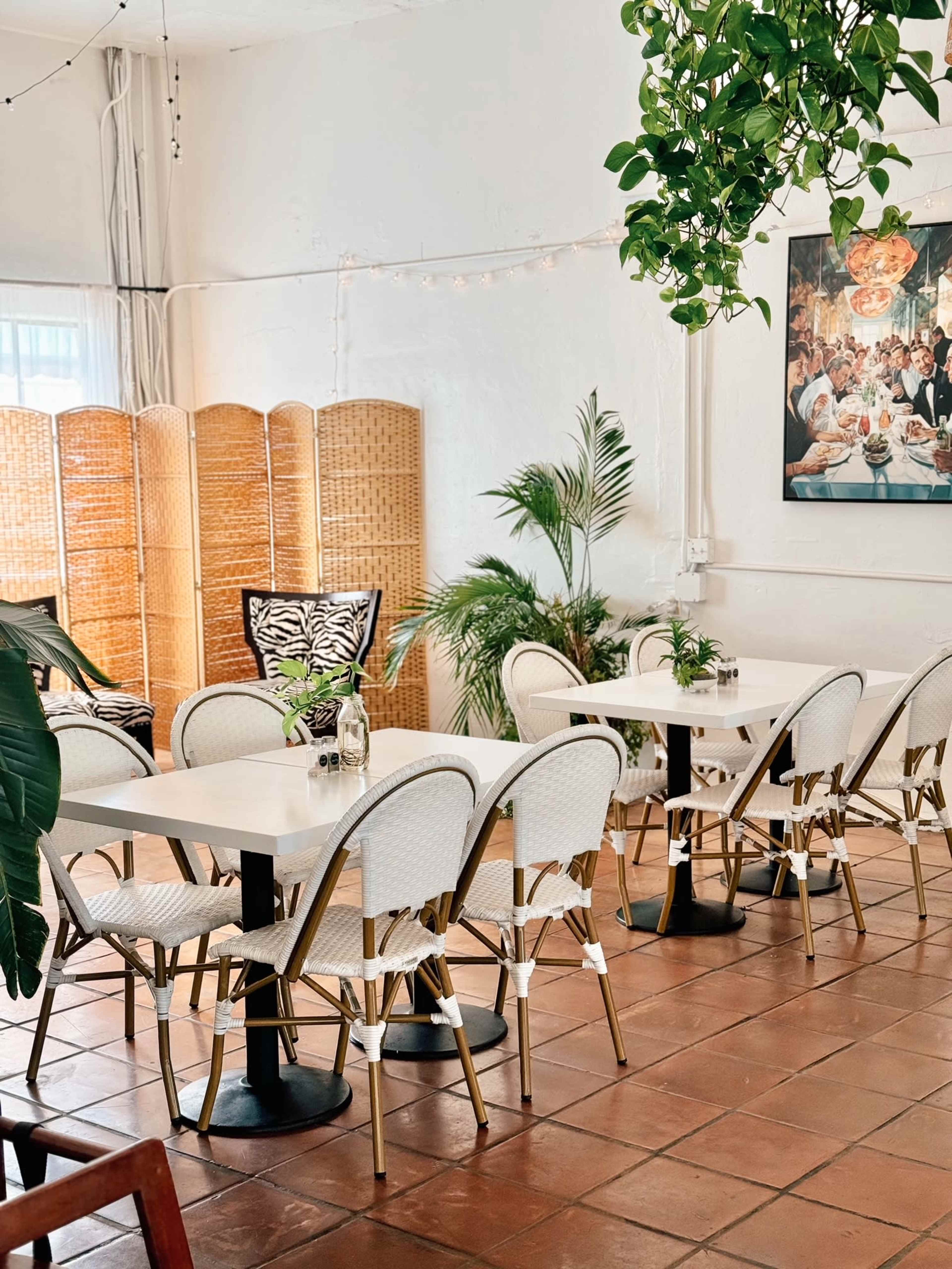 A bright café features several white tables with woven chairs, surrounded by plants and a wooden screen in the background.