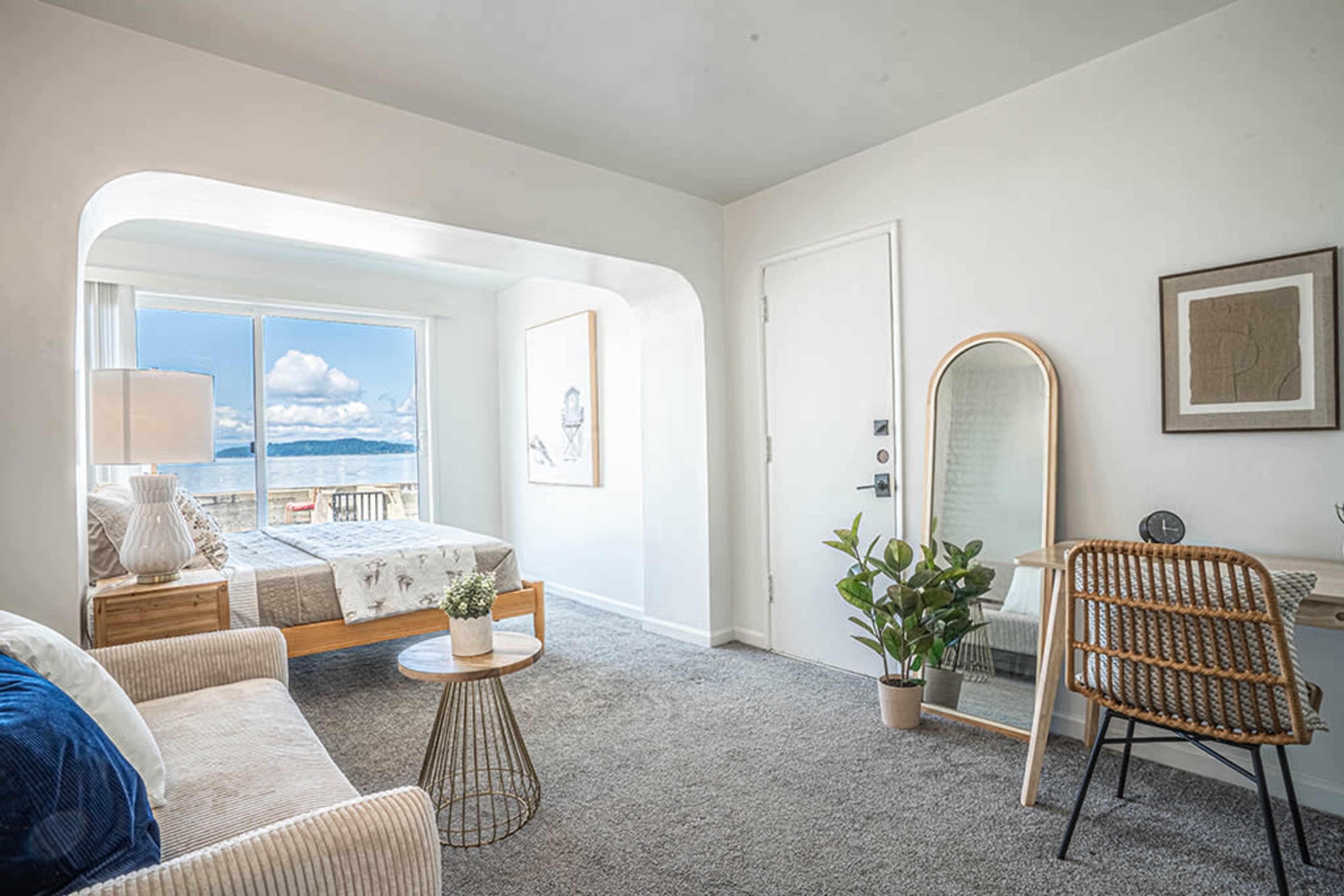 A bright room features a bed, a sitting area with a couch, a desk, and a large window overlooking a scenic view.