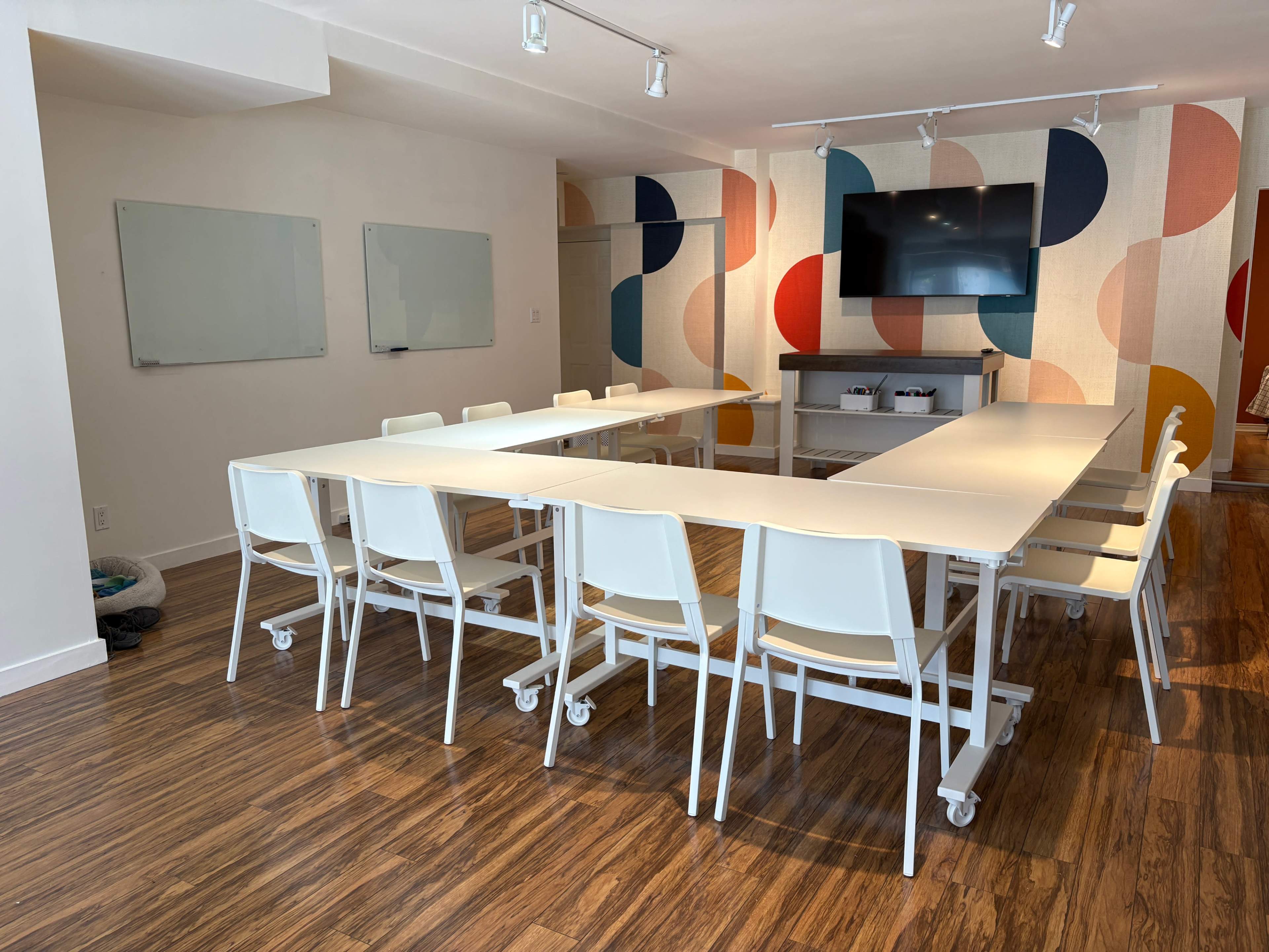 The image shows a modern conference room with a large, elongated table surrounded by white chairs, a wall-mounted television, and colorful patterned panels.