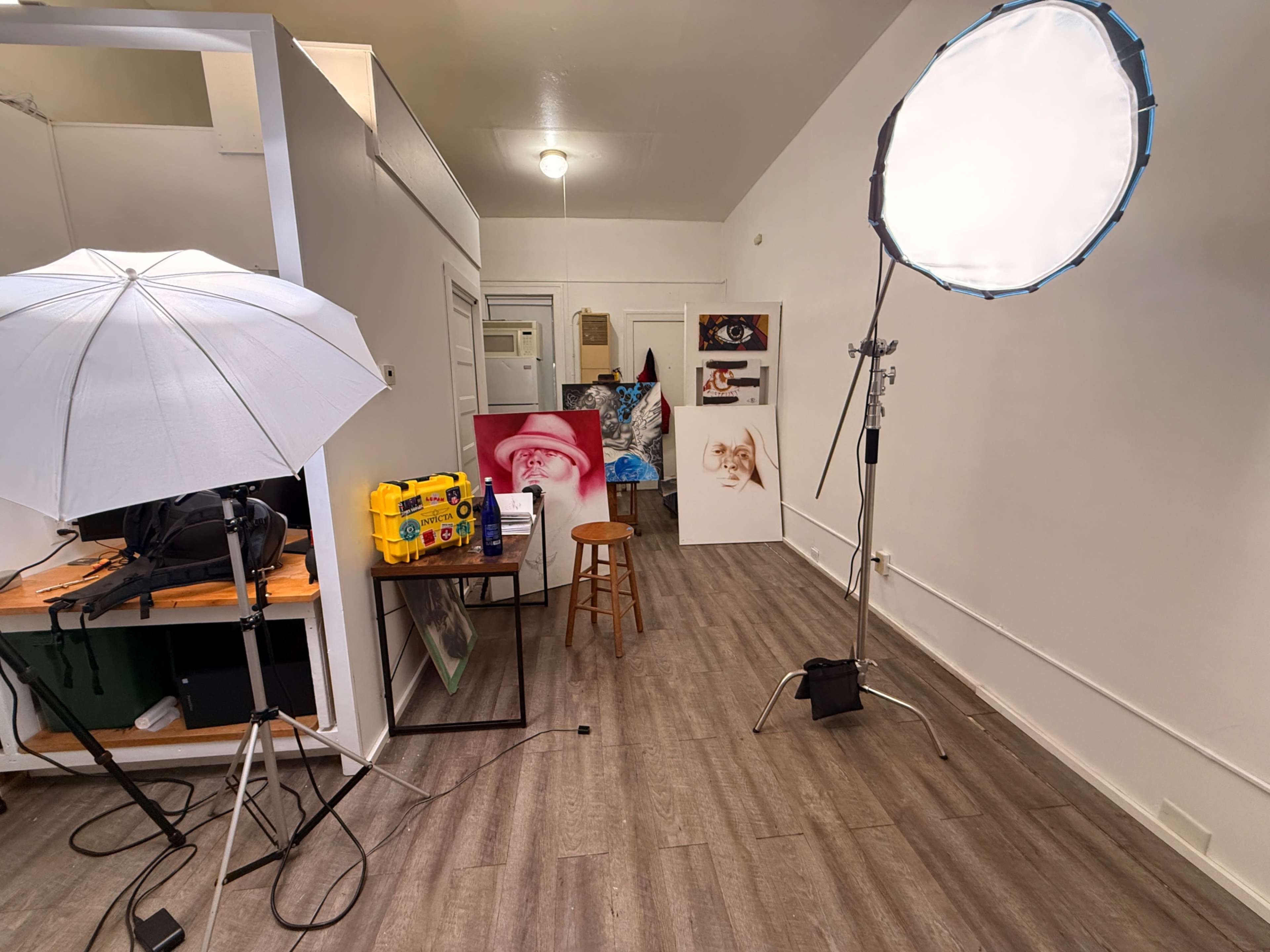 The image shows a well-lit studio space with photography equipment, including umbrellas and a softbox, along with a wooden stool and various printed images on display.