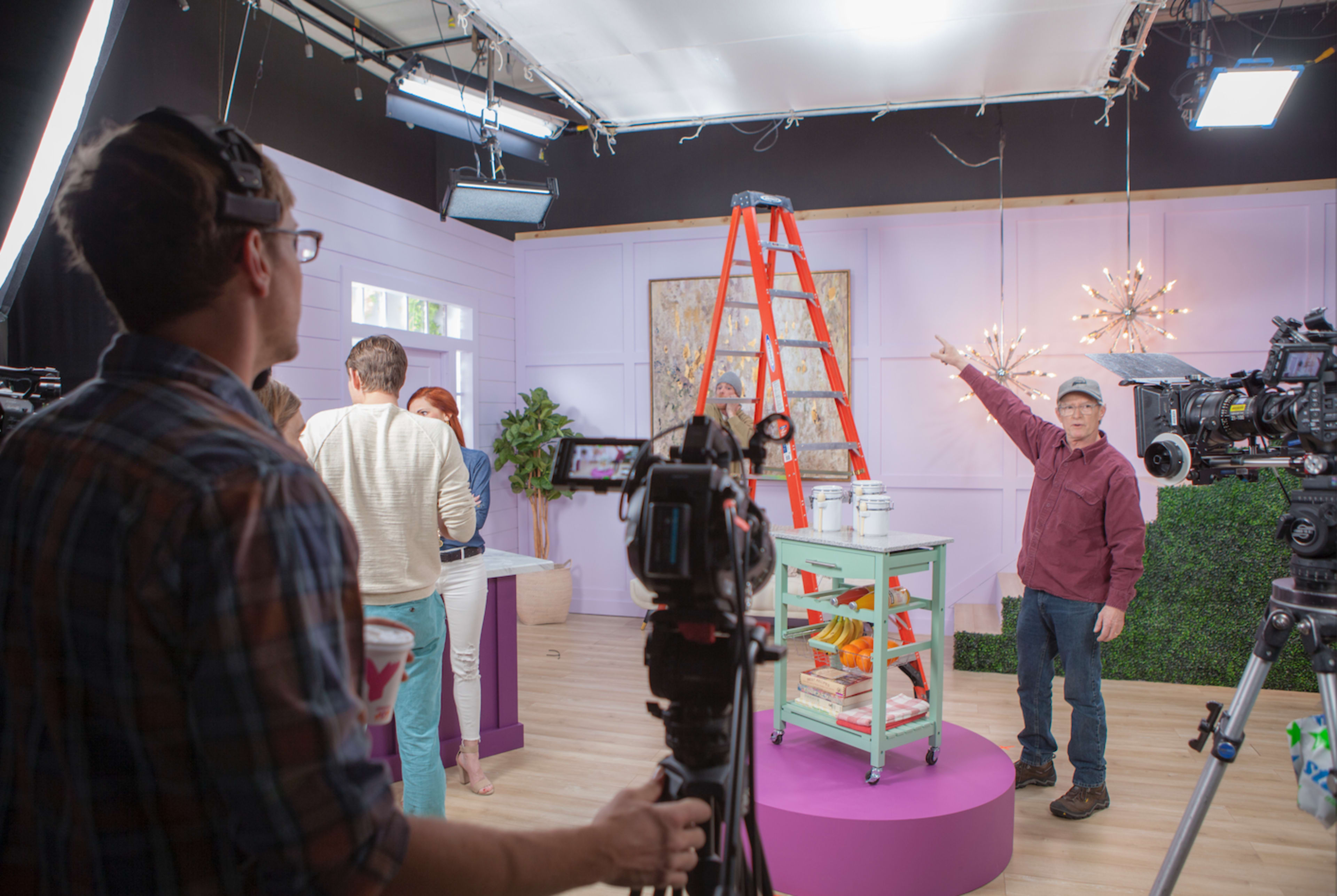 A film crew is set up in a studio space, with a director pointing towards a ladder while cameras and crew members prepare for a shoot.