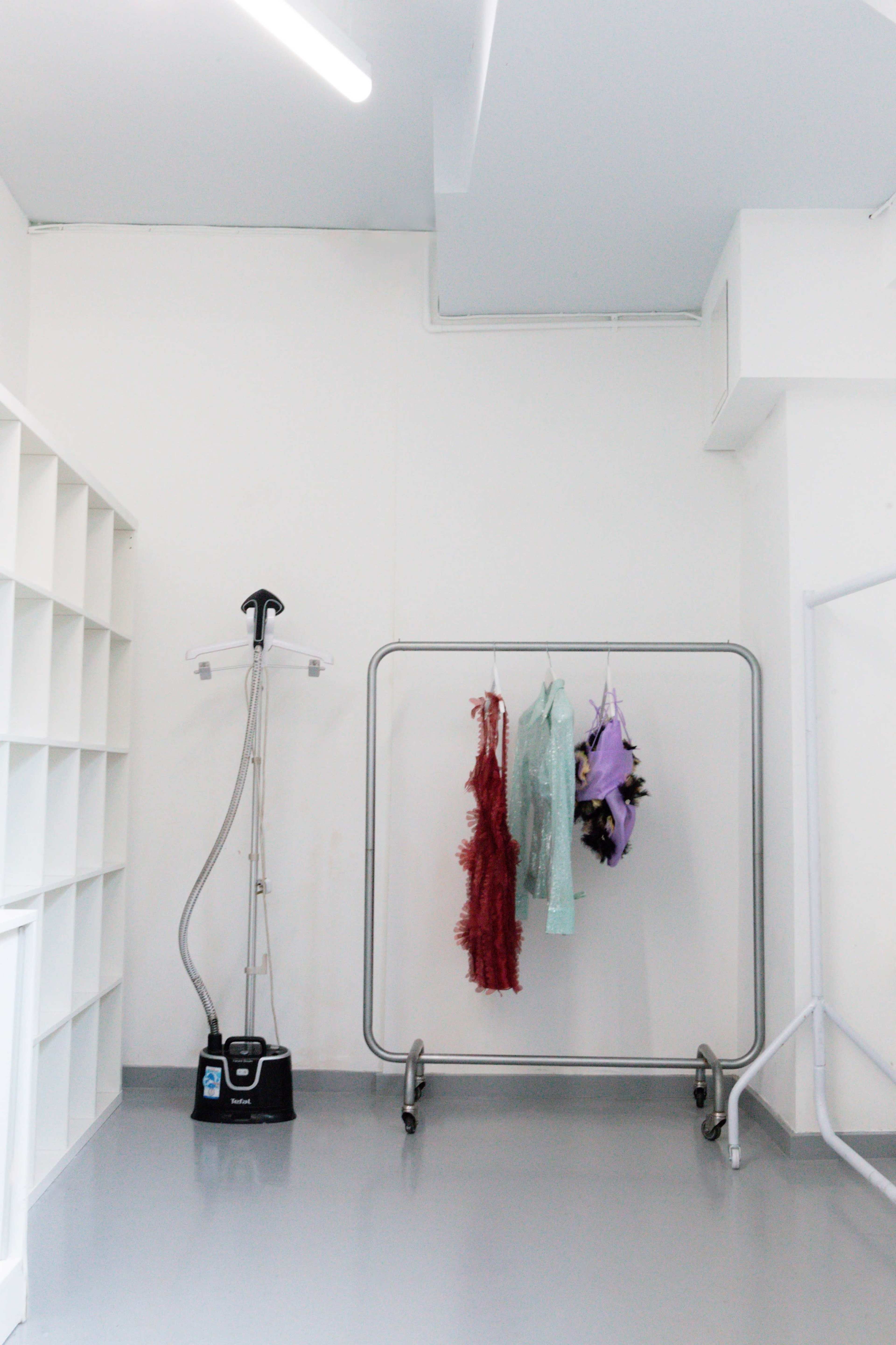 A minimalist room features a steamer on the left and a clothes rack with colorful garments on the right, set against plain white walls.