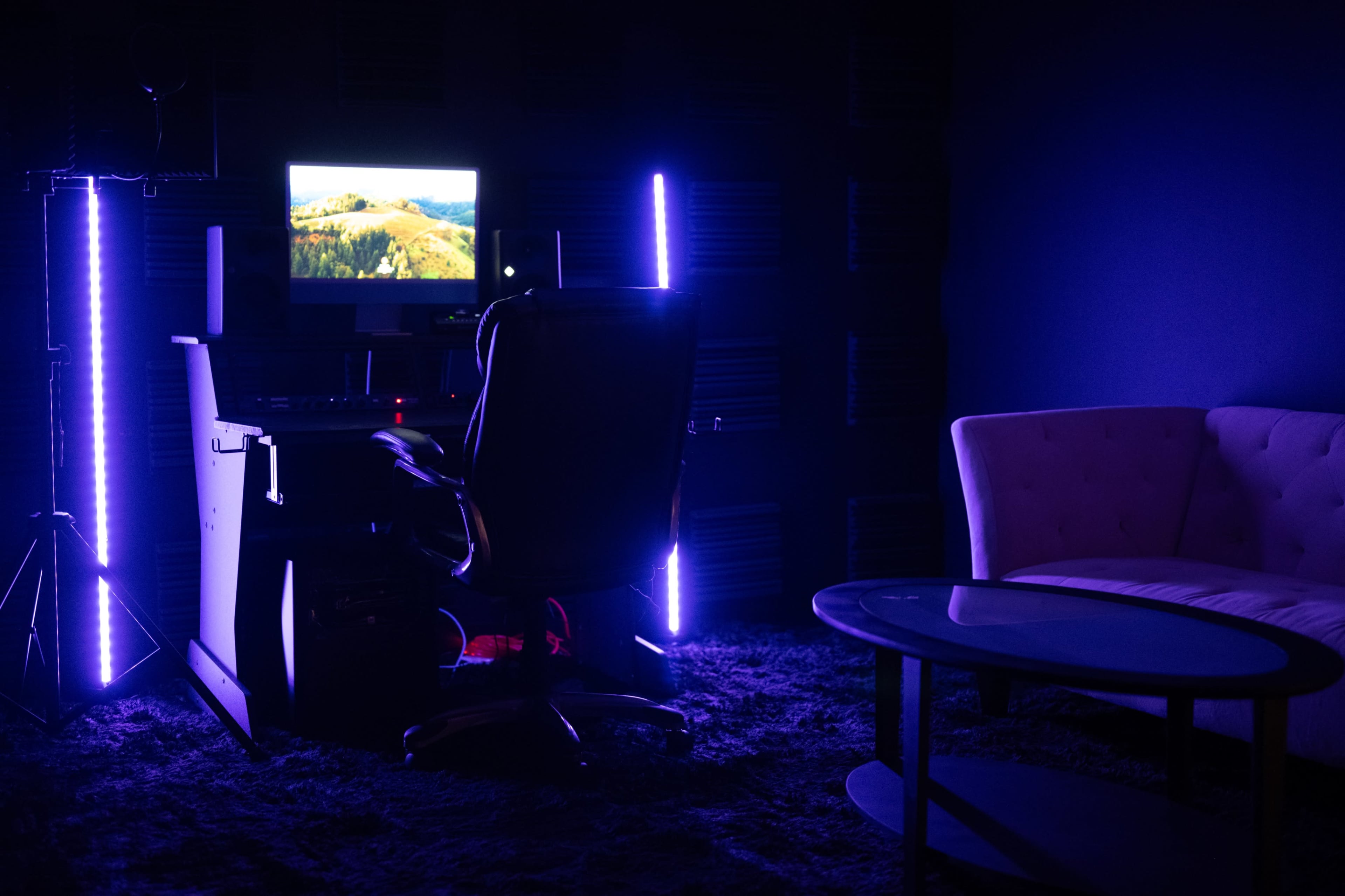 A dimly lit room features a desk with a computer monitor, an office chair, and a pink couch beside a glass coffee table.