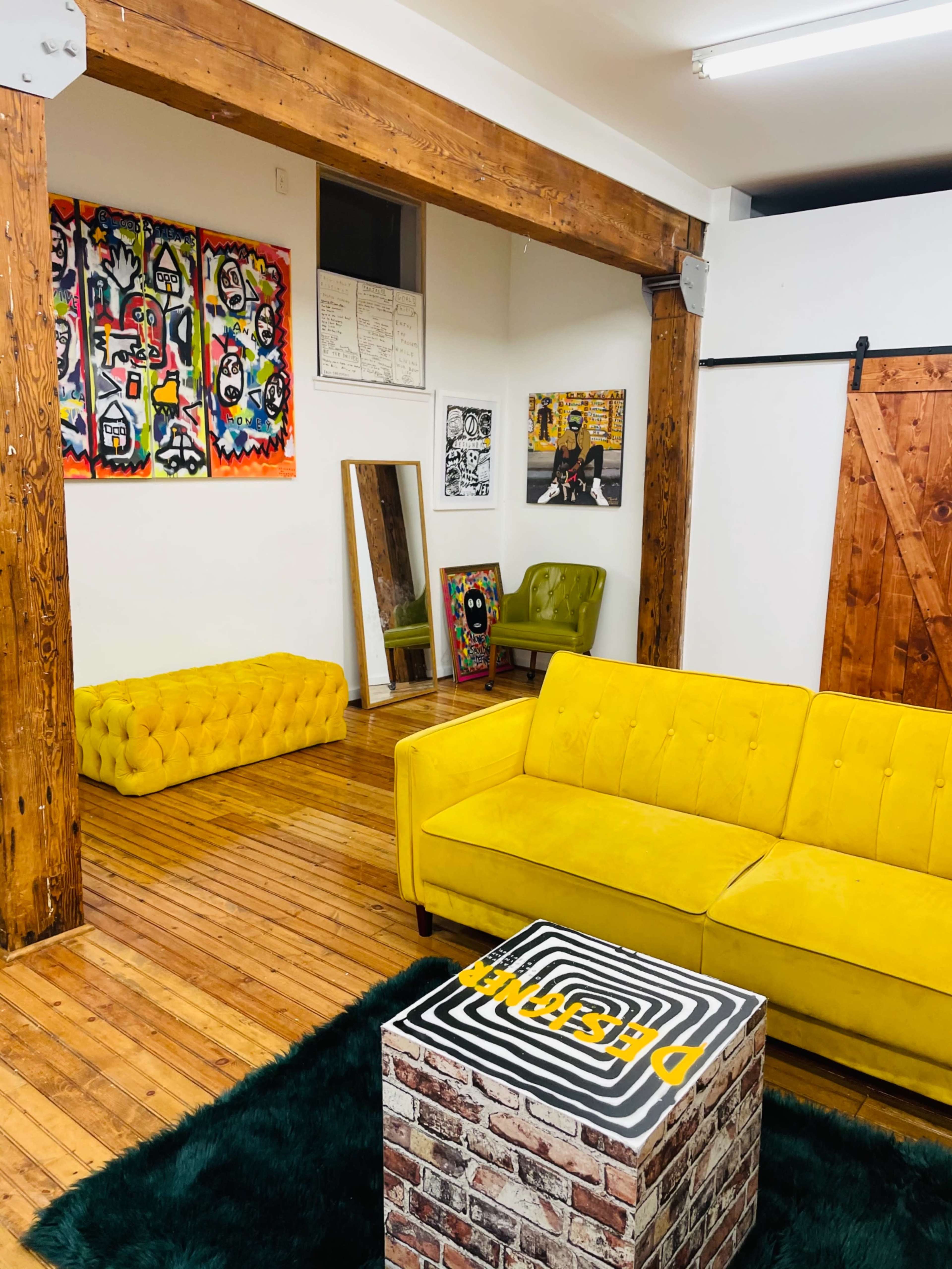 A vibrant room with wooden beams, featuring a yellow couch, a green rug, a brick-patterned coffee table, and various colorful artworks on the walls.