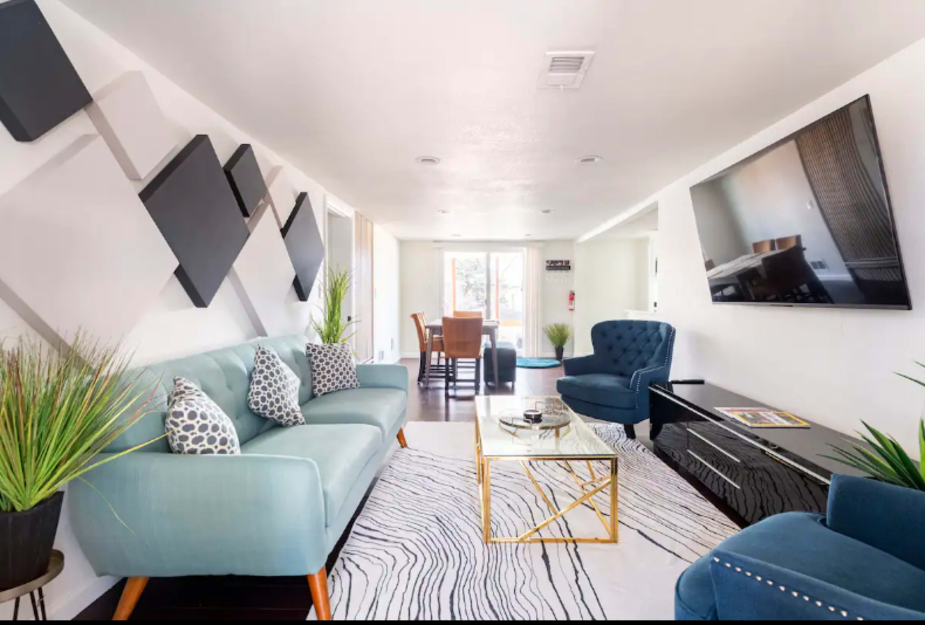 A modern living room features a blue sofa, two blue chairs, a glass coffee table, and a dining area in the background.