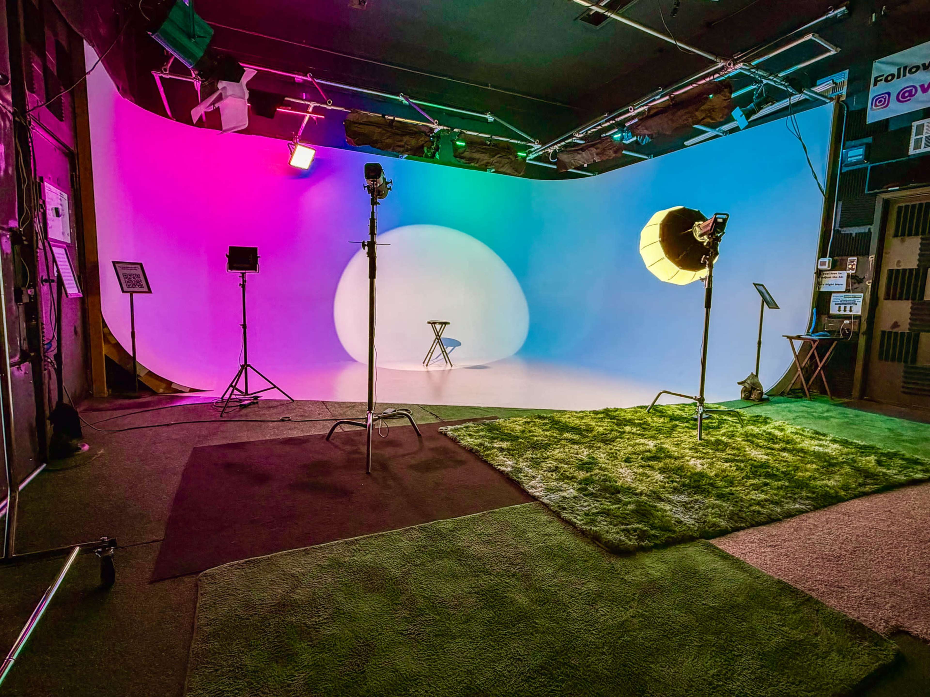 The image shows a photography studio with colorful backdrop lighting, a central white rounded display, and lighting equipment positioned around a small stool.