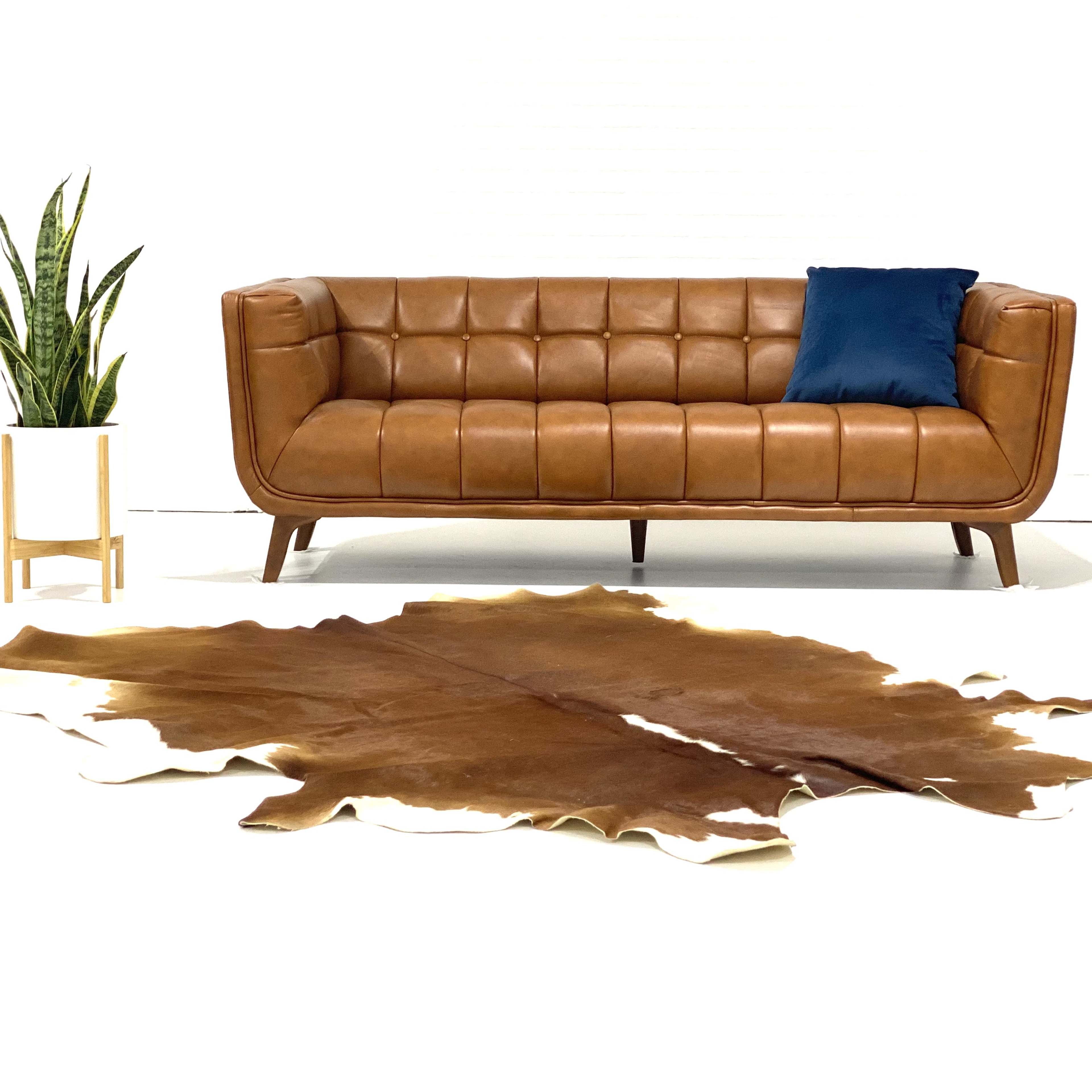 A brown leather sofa with tufted cushions sits on a white floor, accompanied by a potted plant and a cowhide rug.