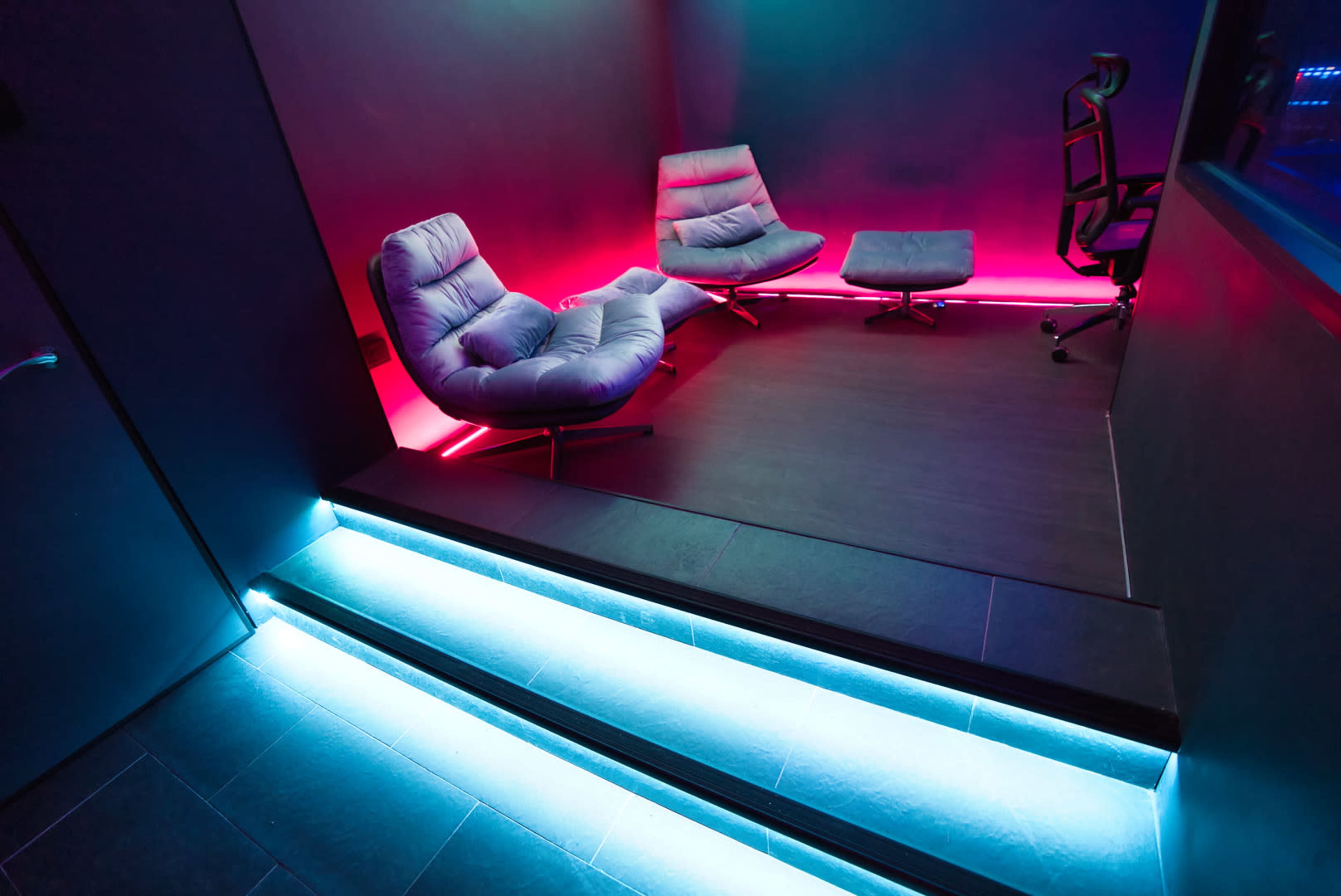 A modern room with two gray lounge chairs and an office chair, illuminated by colored LED lights along the walls and stairs.