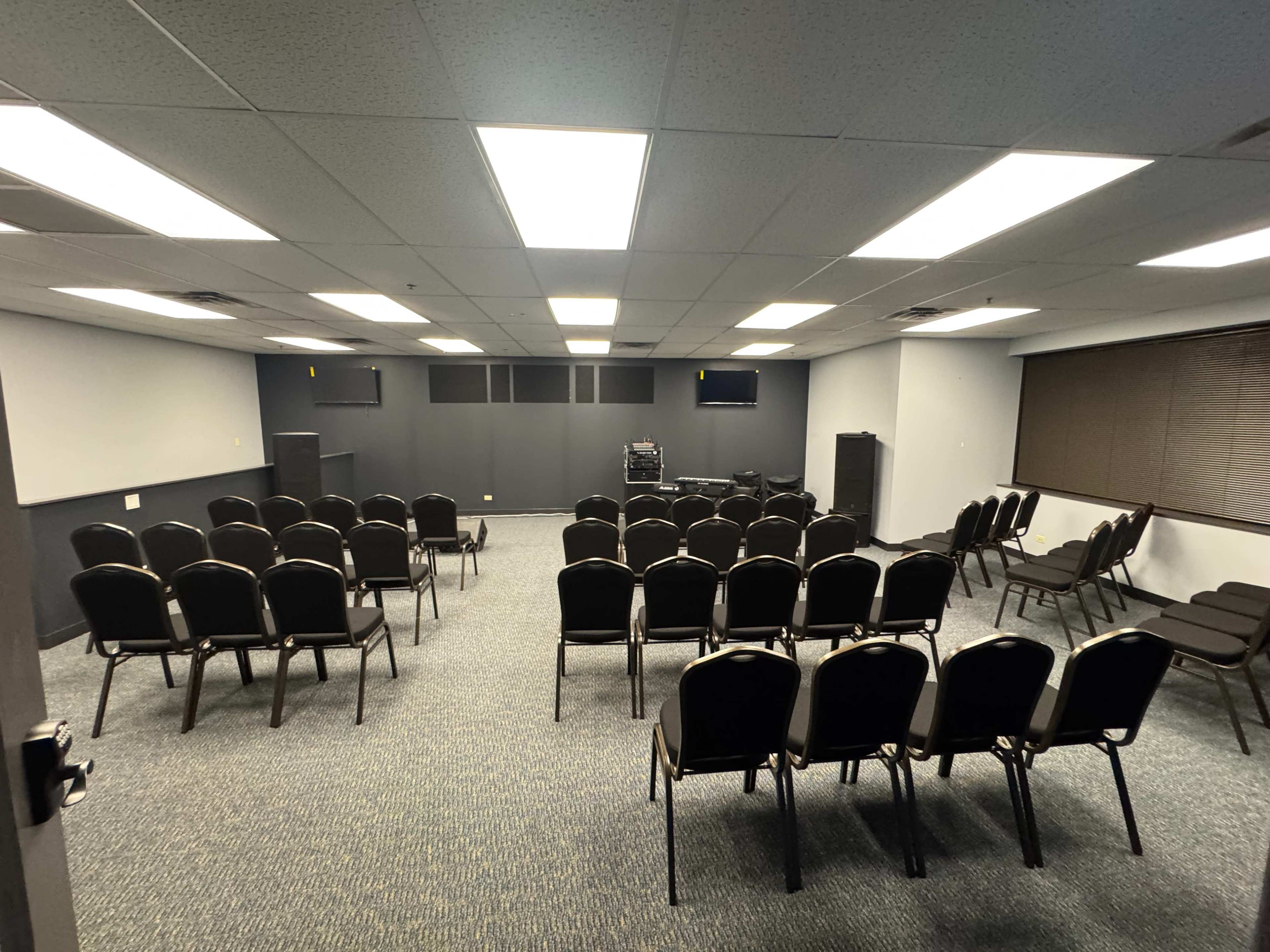 The image shows a conference room arranged with multiple rows of black chairs facing a projection screen, lit by bright overhead lights.