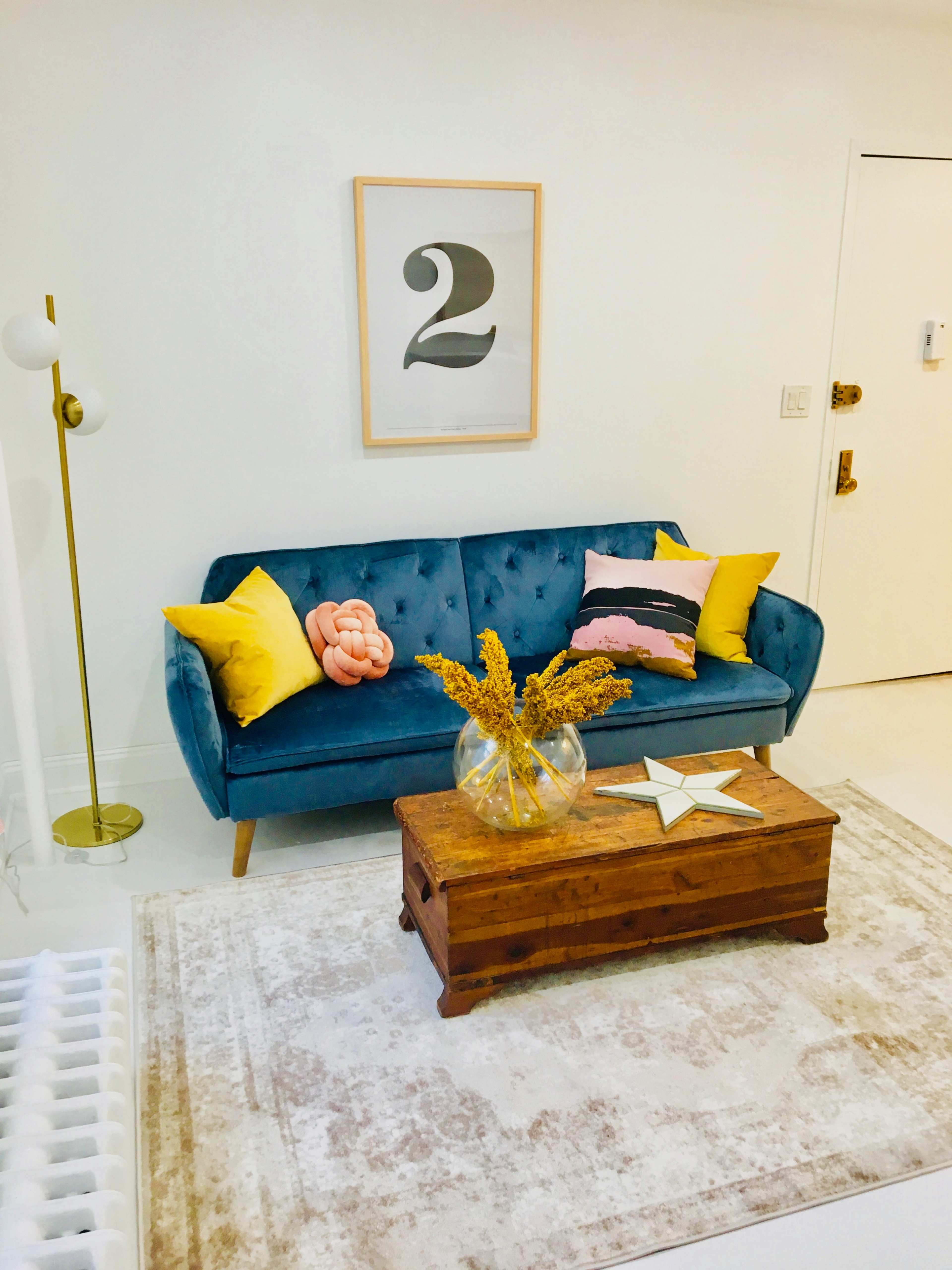 A blue couch with yellow and pink accent pillows sits in a bright room featuring a wooden coffee table and a vase of dried flowers.