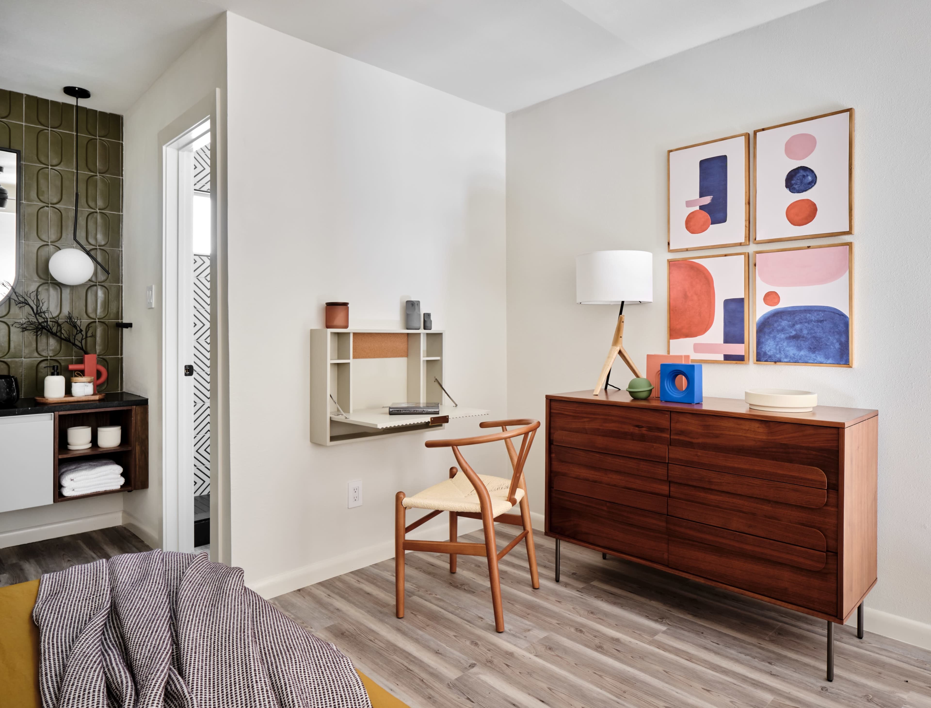 The image shows a modern, well-decorated room featuring a wooden dresser, a chair, a wall-mounted desk, and artwork arranged above the dresser.